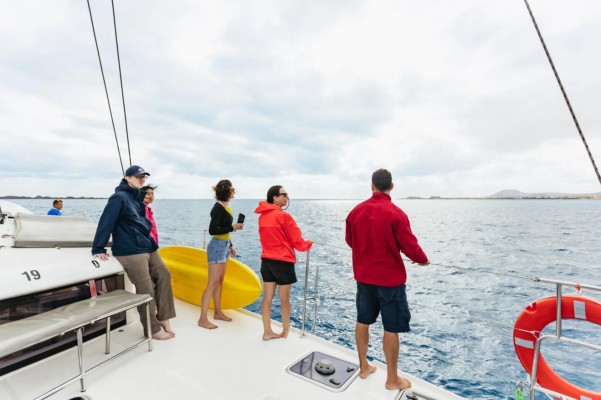 Guests in light jackets at the rail of a catamaran under way, with Fuerteventura on the horizon
