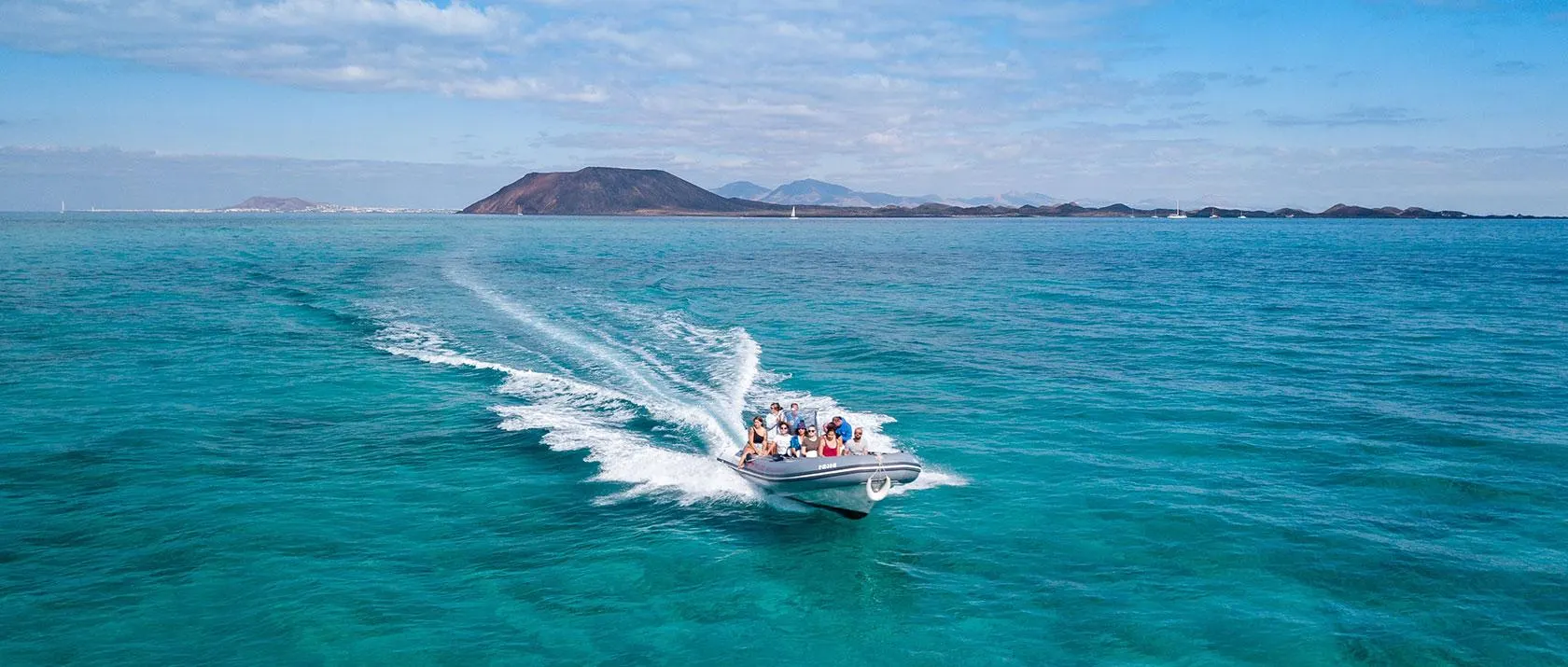 A grey rigid inflatable boat full of passengers speeding across turquoise water, with Isla de Lobos and its volcanic cone behind