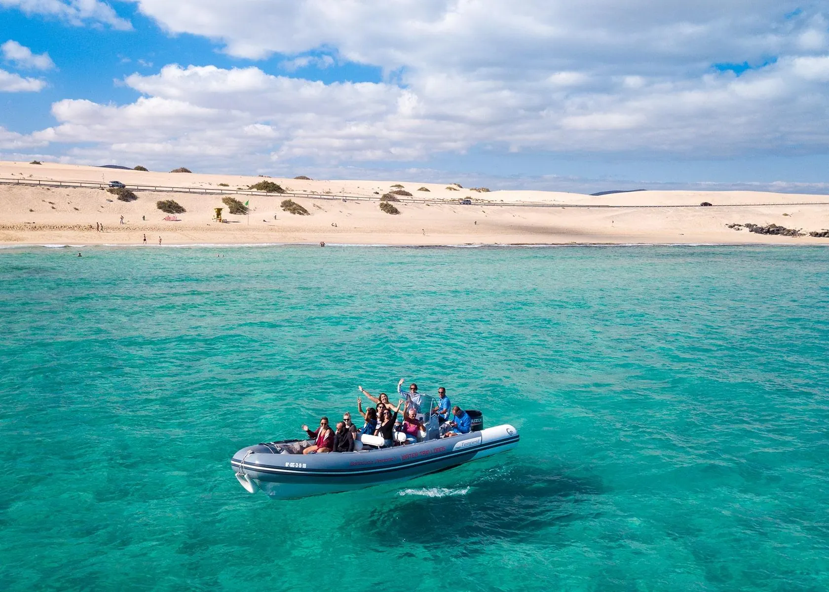 Passengers waving from a Water Taxi Lobos inflatable boat in clear green water off the sand dunes of Corralejo