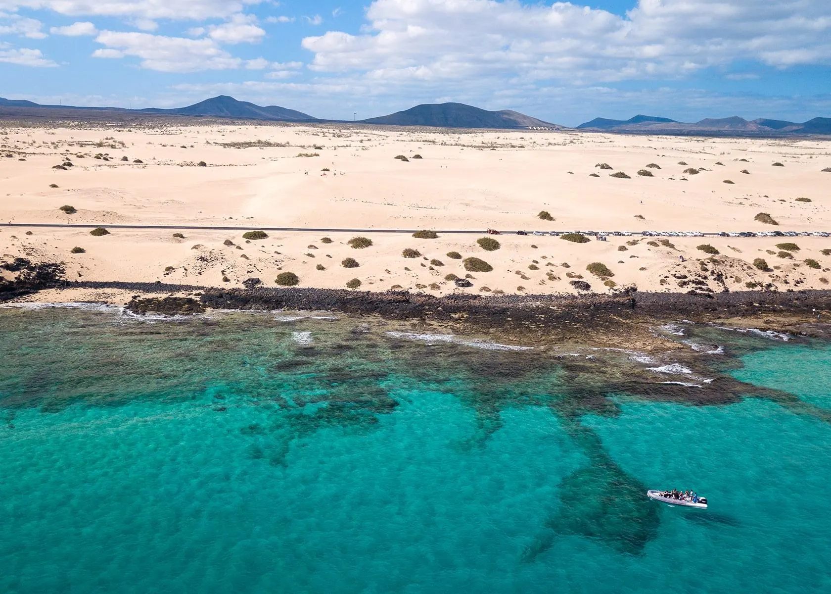 An aerial view of a small boat over reefs off the Corralejo dunes, with volcanic hills on the horizon