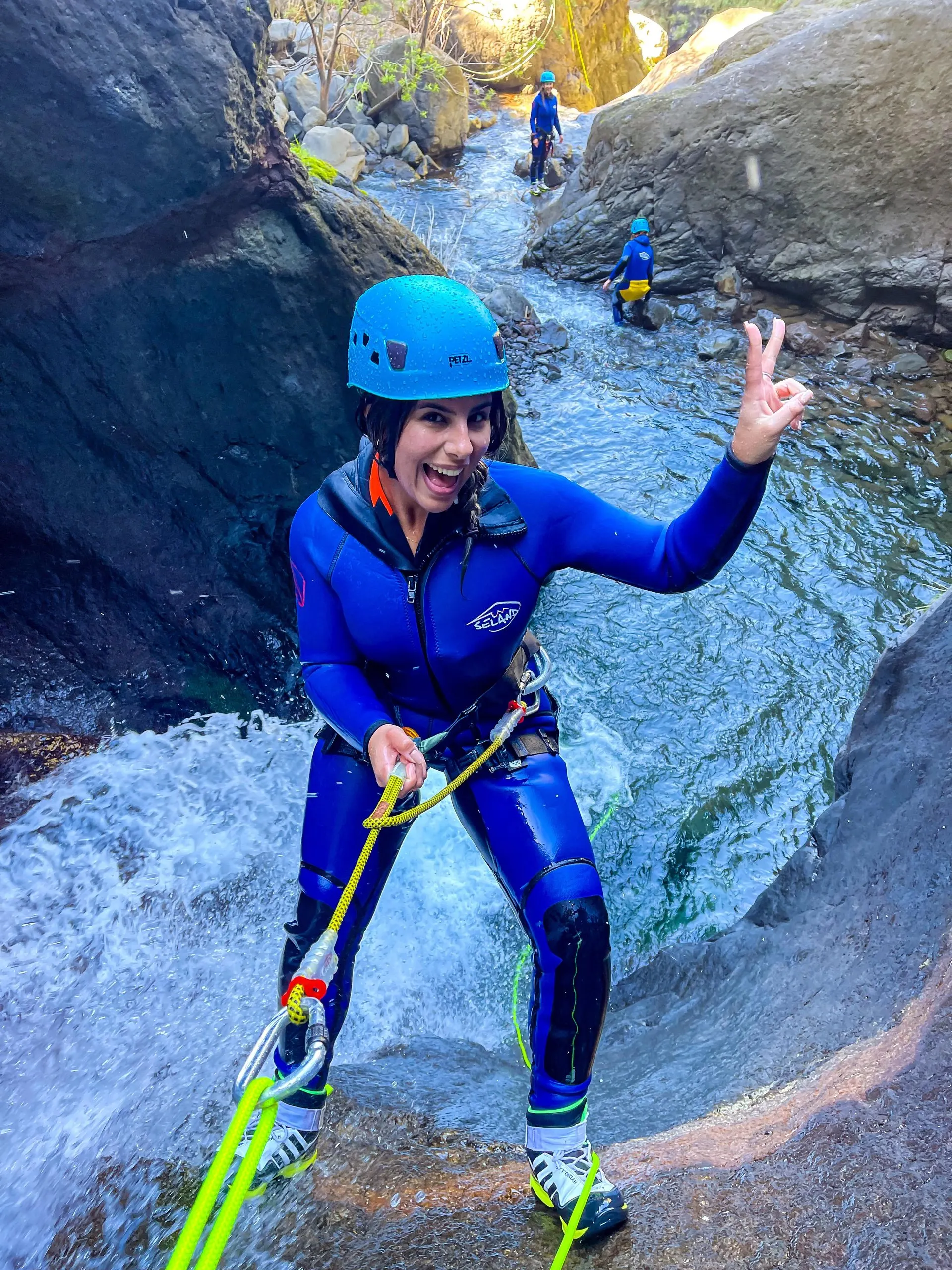 A smiling canyoner in a blue wetsuit and helmet abseiling down a small waterfall on a rope, with two others waiting in the stream below