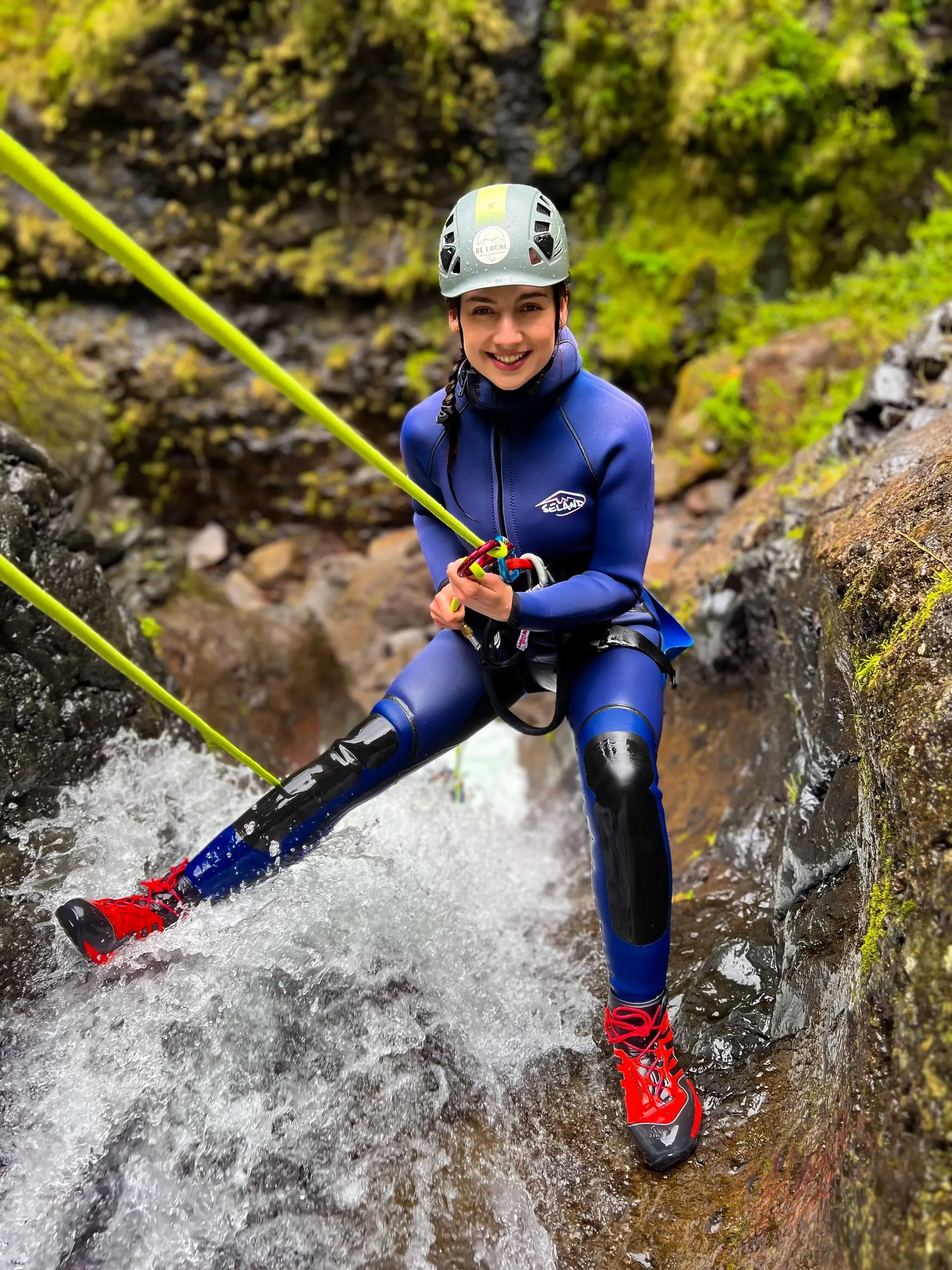 A canyoner in a wetsuit, harness and helmet leaning back on the rope at the top of a waterfall, with mossy rock behind