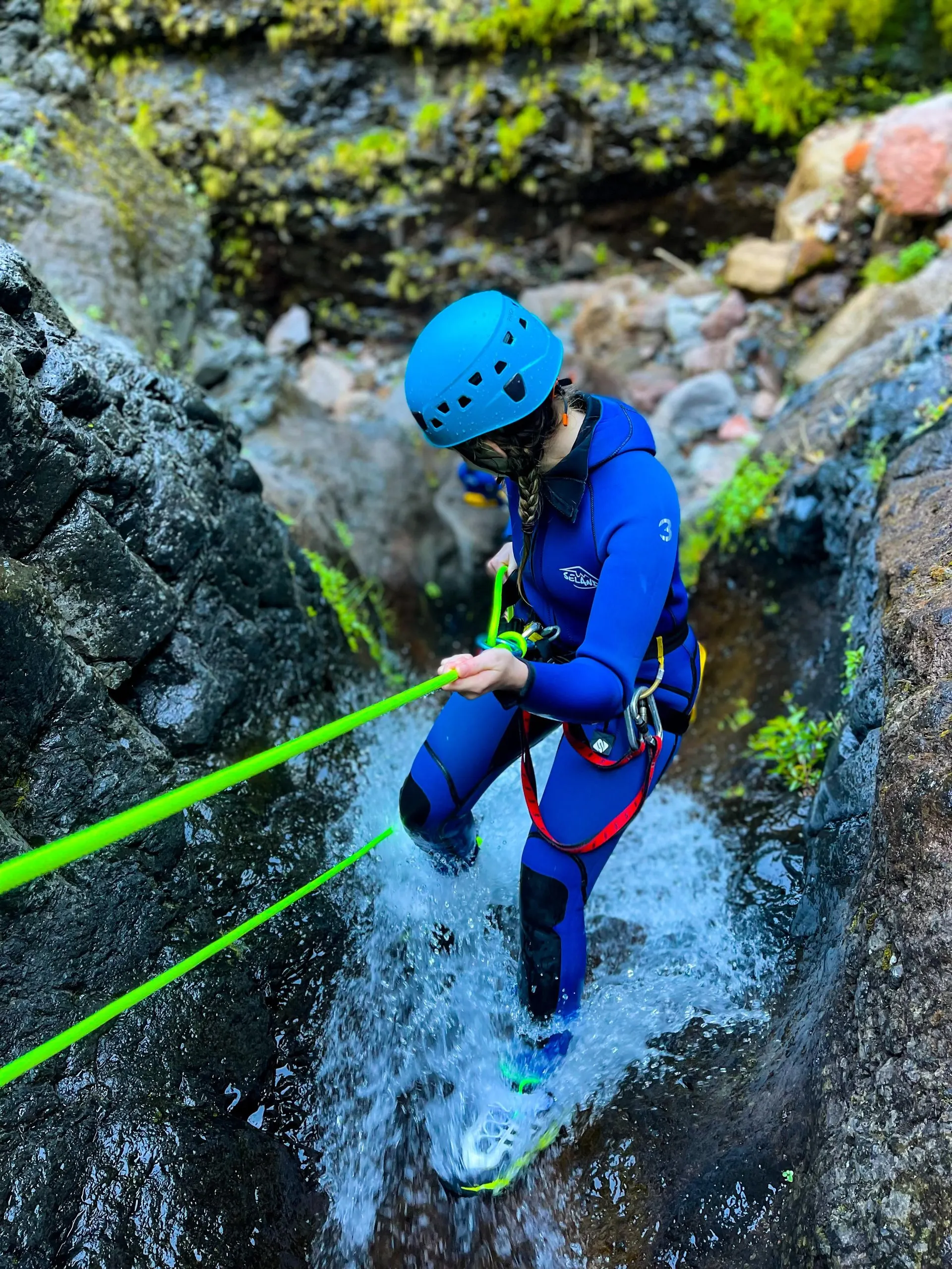 Looking down on a canyoner lowering herself on two green ropes through a narrow waterfall chute of dark volcanic rock