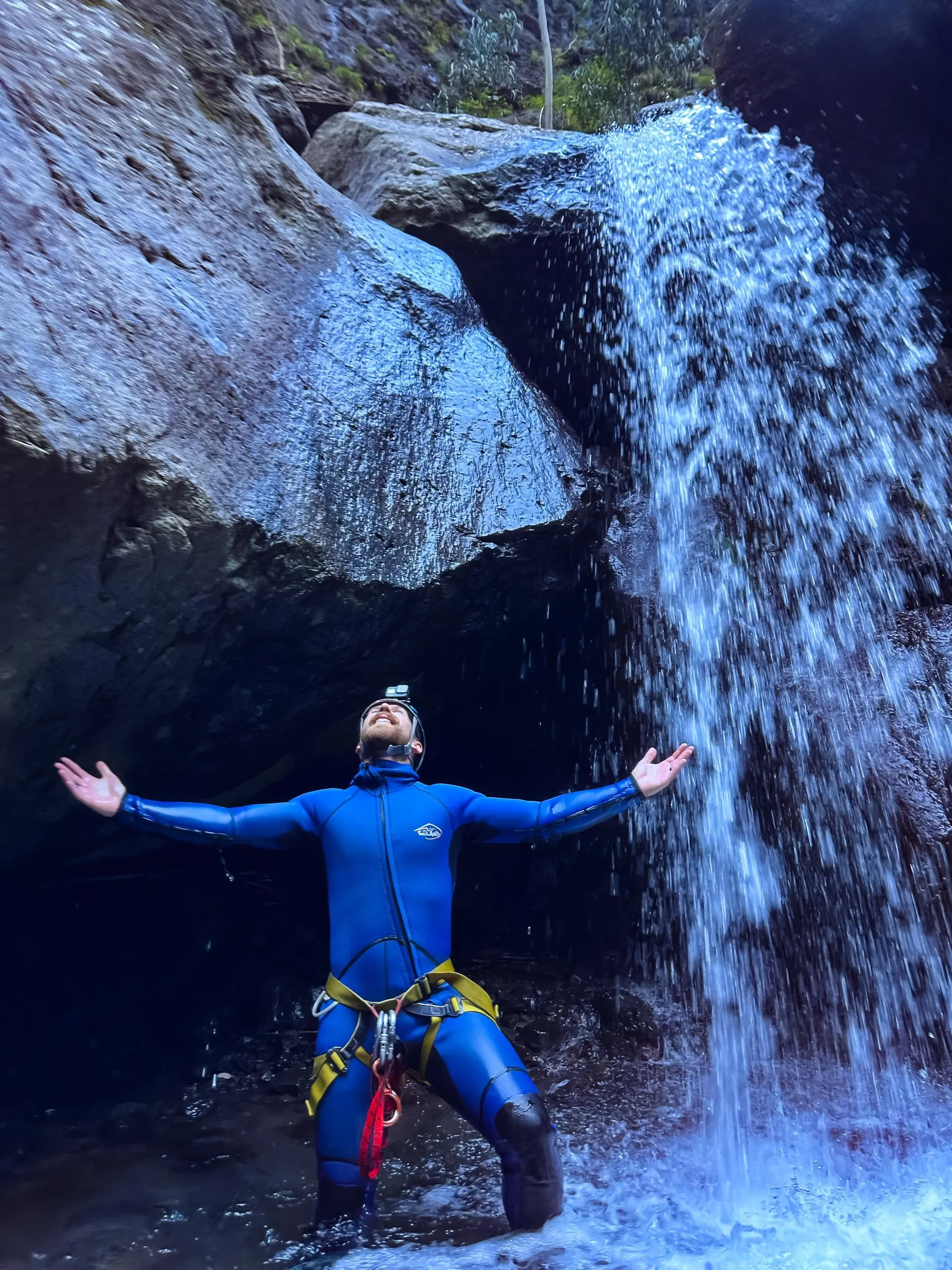 A canyoner standing in a pool at the foot of a waterfall under a huge boulder, arms spread wide