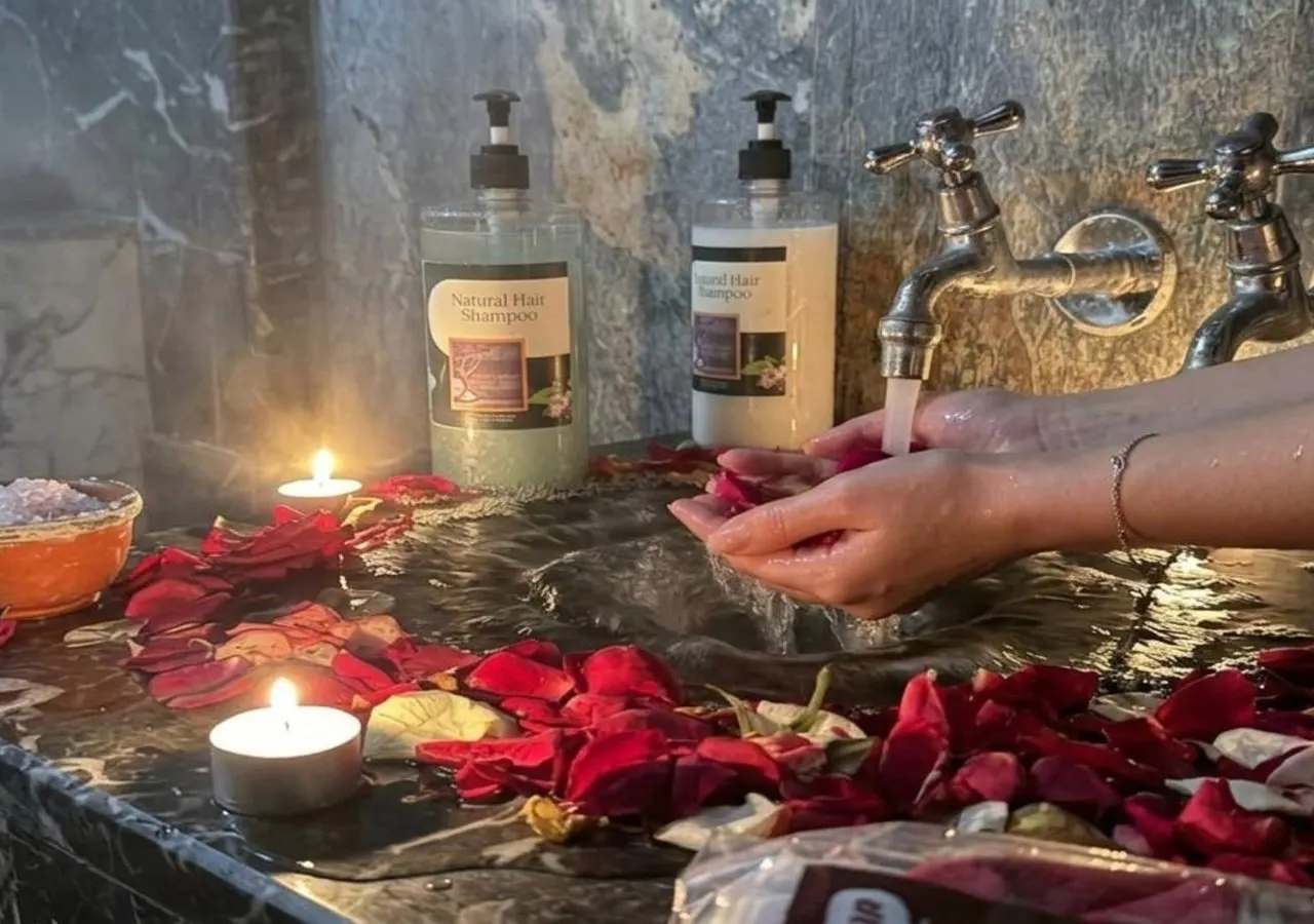 Hands rinsing rose petals under a tap in a marble hammam, with candles and bottles of natural shampoo