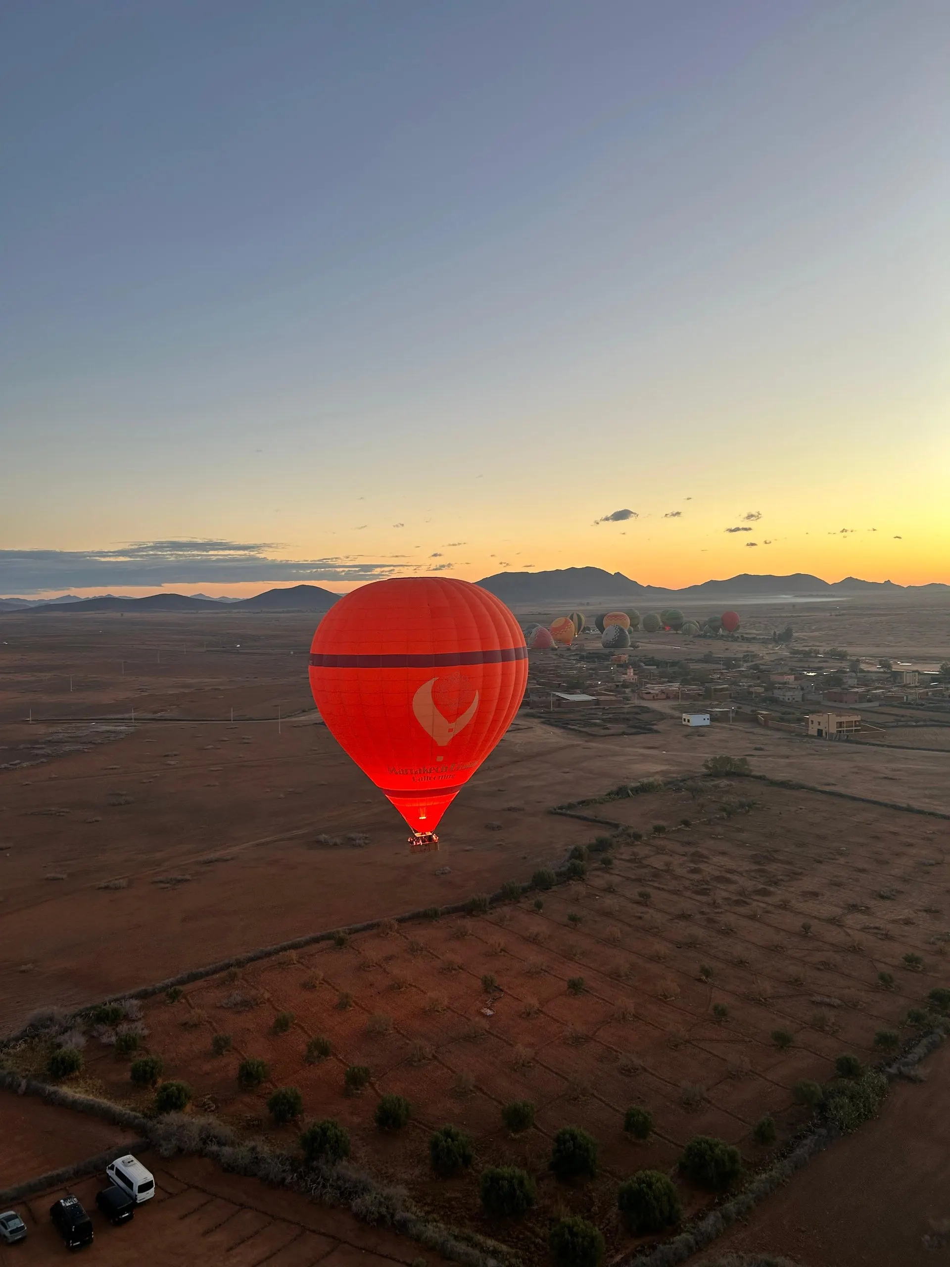 A red Marrakech Dream Ballooning balloon in flight at dawn over red farmland and a village, with other balloons inflating and low hills on the horizon