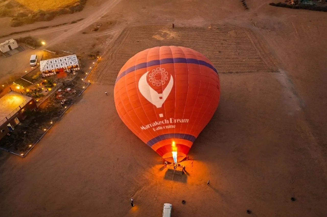 Looking down on an orange Marrakech Dream Ballooning balloon with its burner lit at the take-off field, beside the lit breakfast tents and a minibus