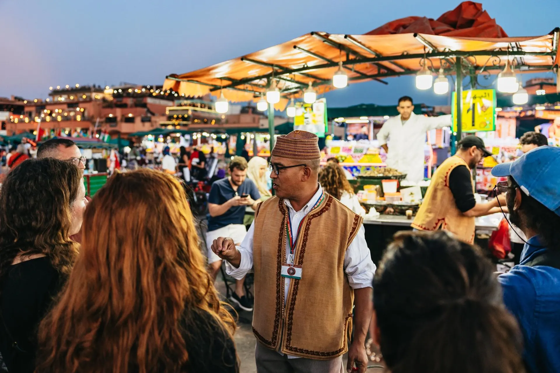 A guide in a waistcoat and cap talking to his group among the lit food stalls of Jemaa el-Fna at dusk