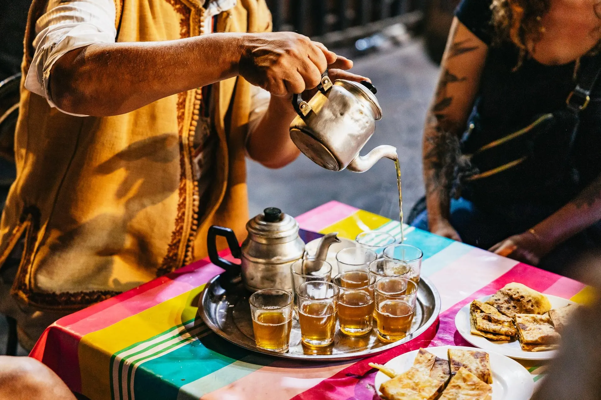 Mint tea poured from a metal teapot into glasses on a striped tablecloth, with plates of msemen flatbread