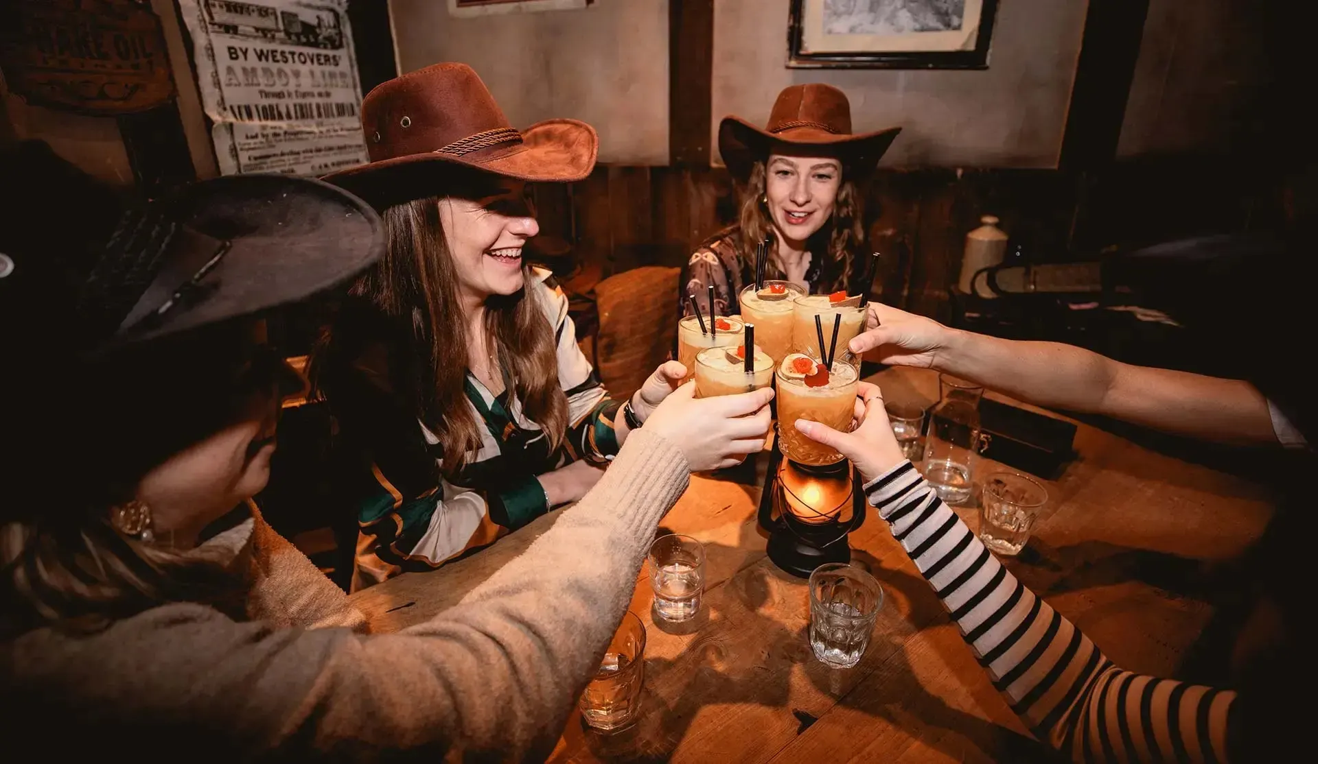 Friends in cowboy hats clink tall orange cocktails over a wooden saloon table lit by a lantern