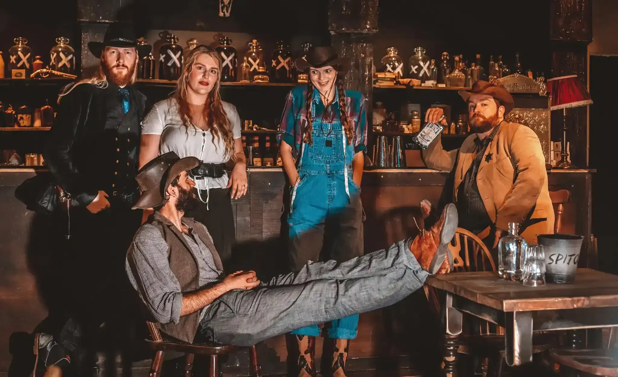 Five actors in Wild West costume pose in front of shelves of moonshine jars, one leaning back with his boots on the table