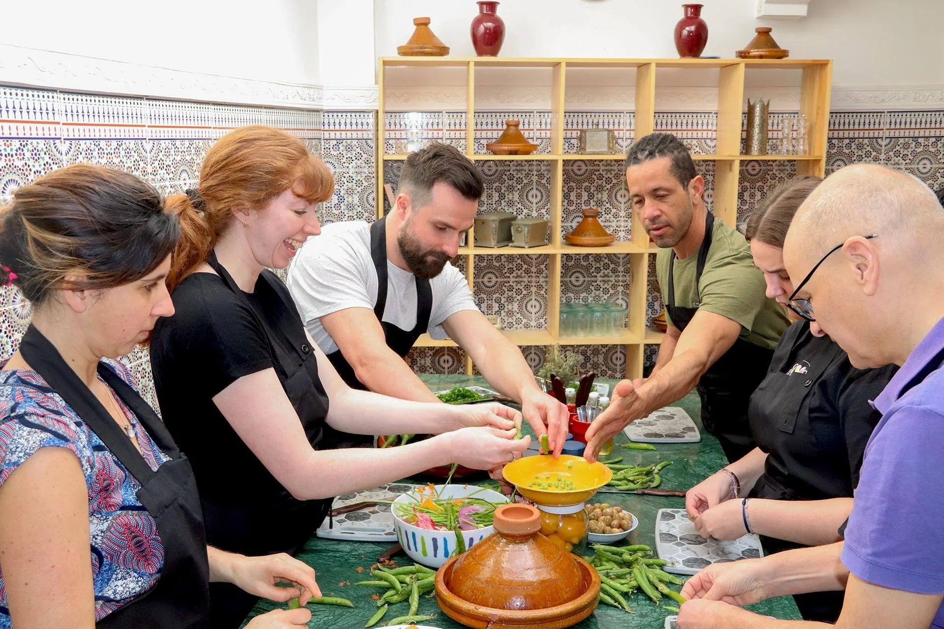 Guests in aprons preparing vegetables around a tiled kitchen table, with a clay tagine in front of them