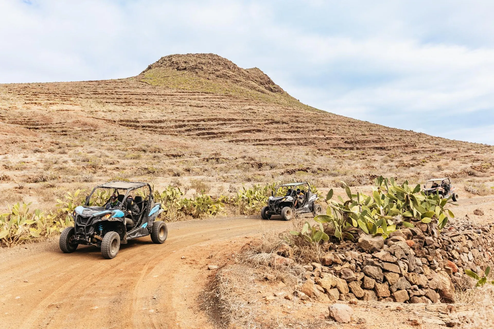 Three two-seat buggies on a dirt track lined with prickly pear cactus, below a terraced volcanic hill in northern Lanzarote