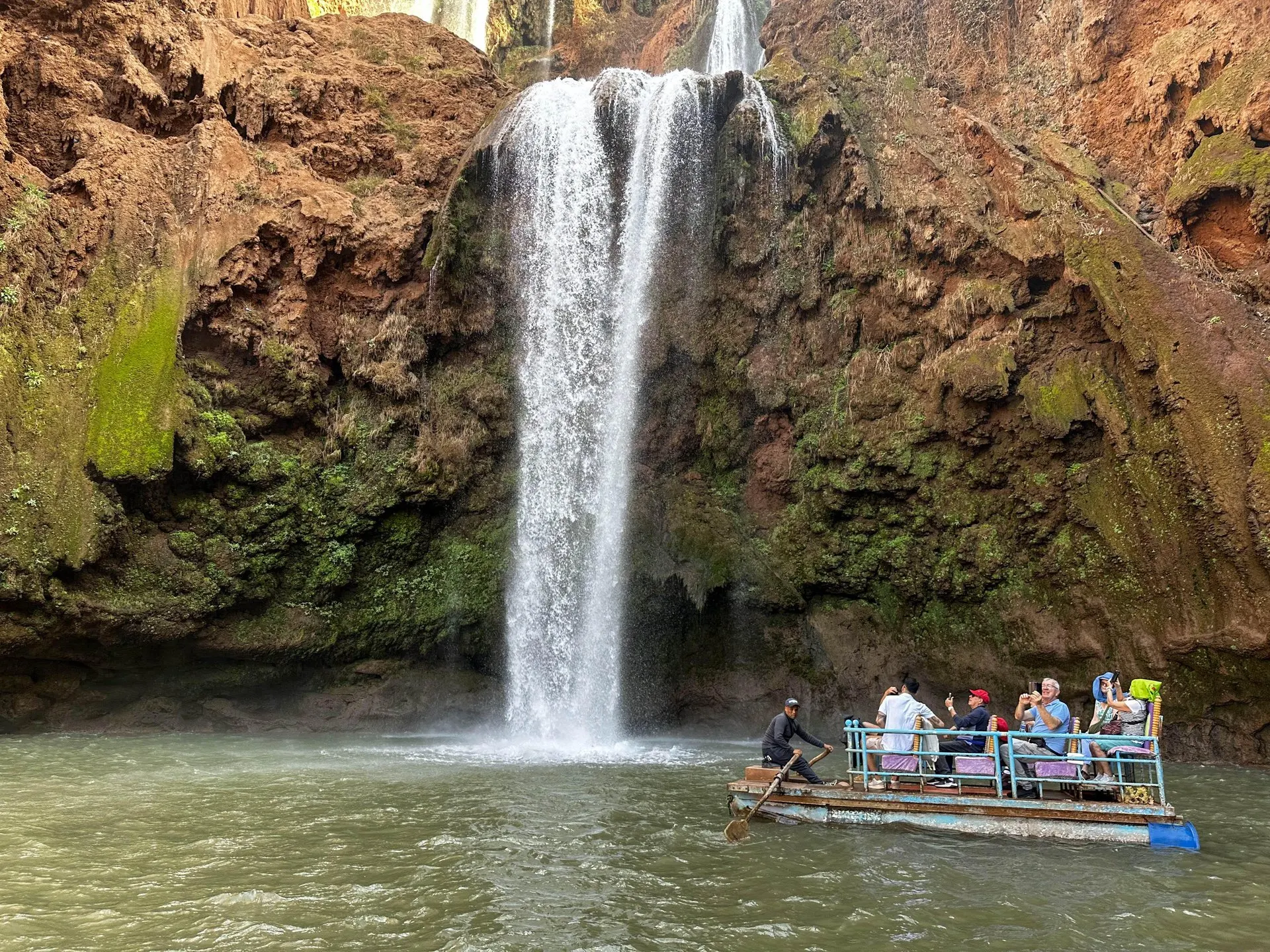 A wooden boat of visitors, rowed by a boatman, in the pool at the foot of one of the Ouzoud waterfalls, below red rock and moss