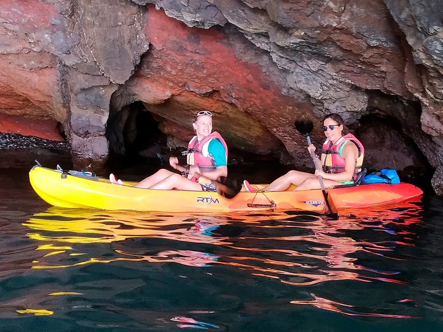 Two paddlers in a double kayak at the mouth of a sea cave in red and grey volcanic rock