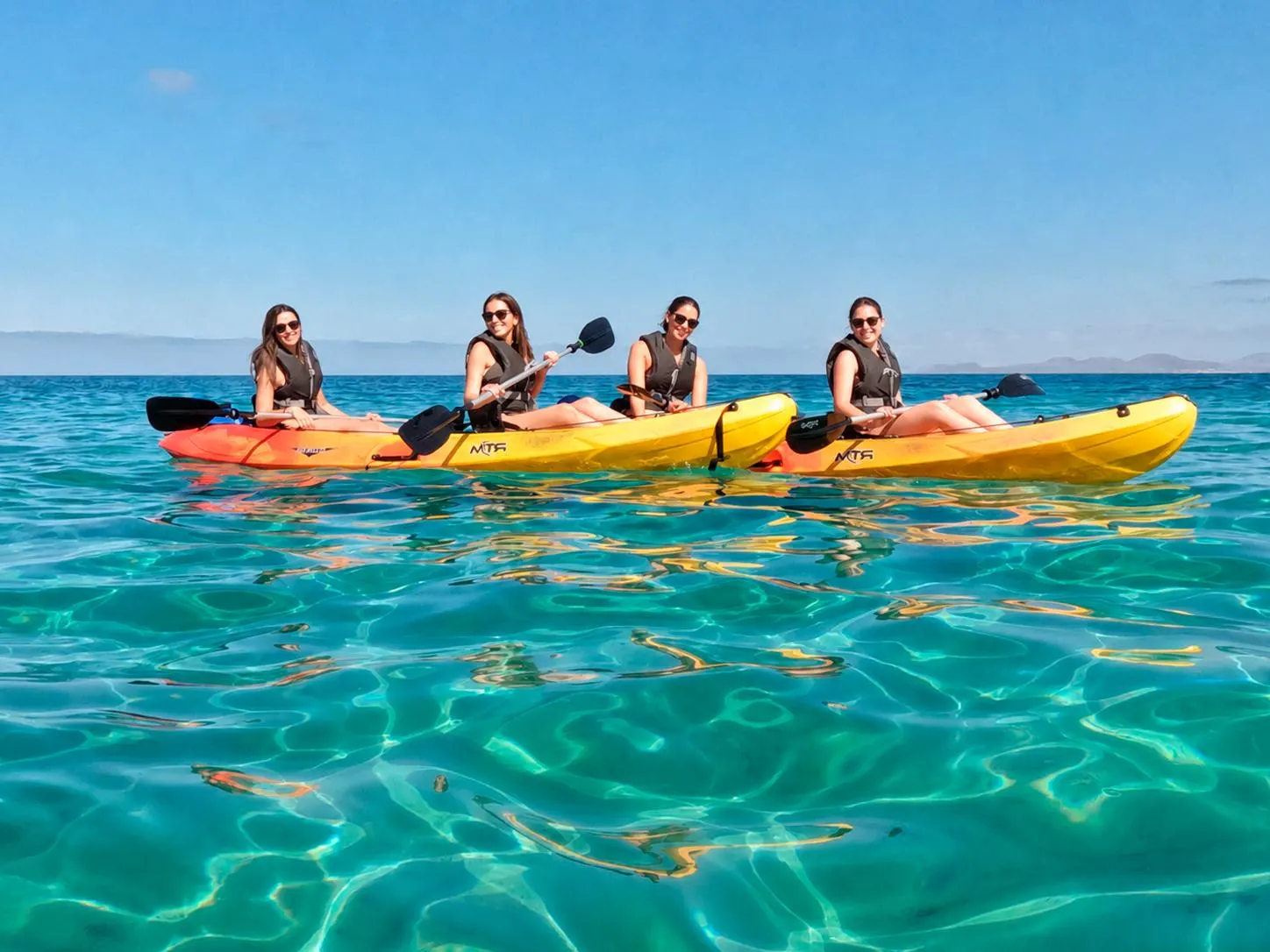 Four paddlers in two double kayaks on calm, clear water, with the coast of Lanzarote low on the horizon