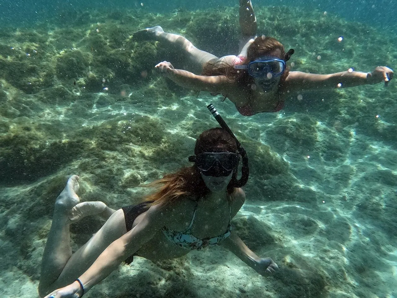 Two snorkellers in masks swimming over a sandy, rocky seabed in clear water