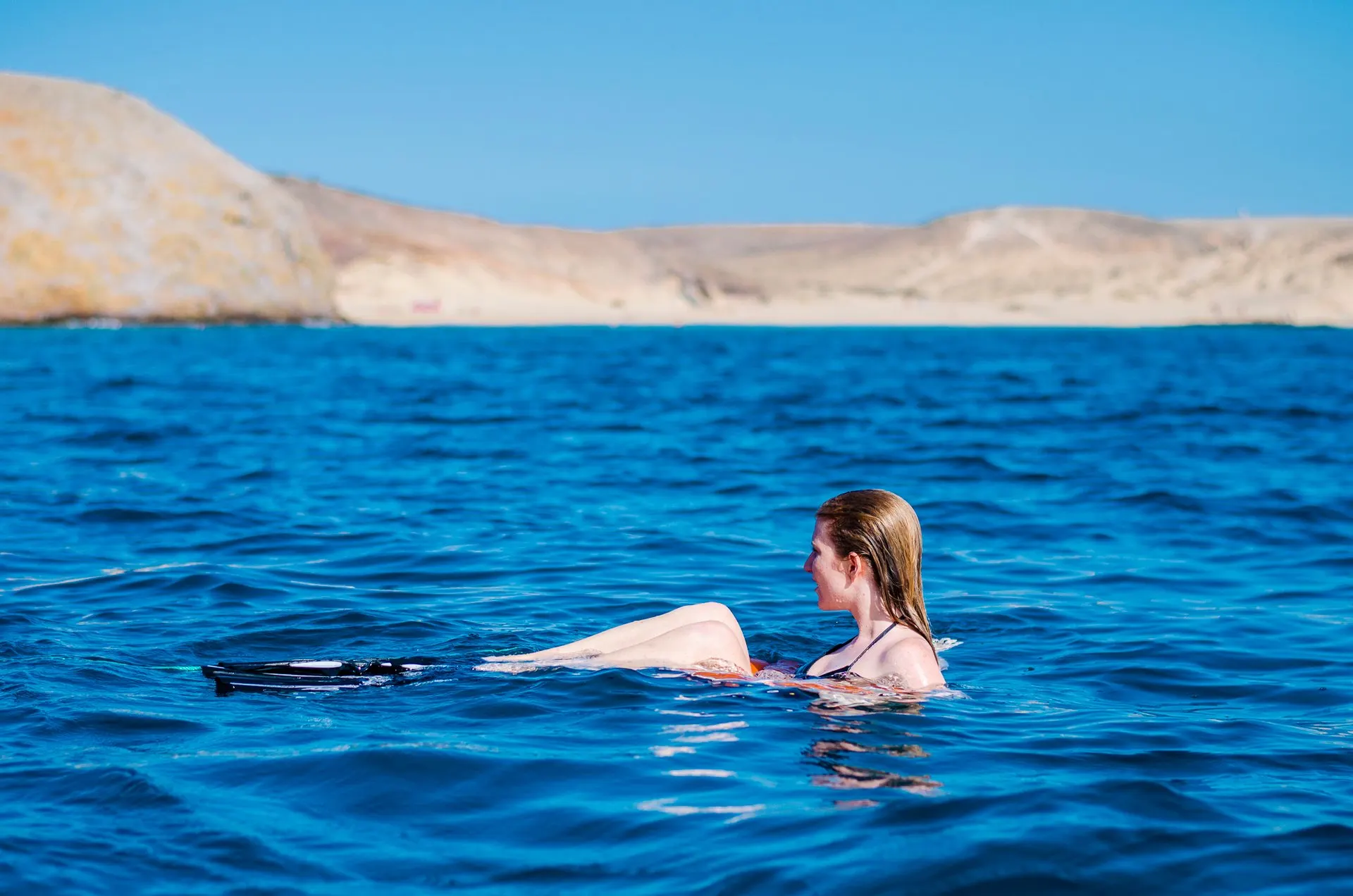 A swimmer with snorkelling fins in deep blue water off Papagayo, with the beach and the bare hills of Los Ajaches behind