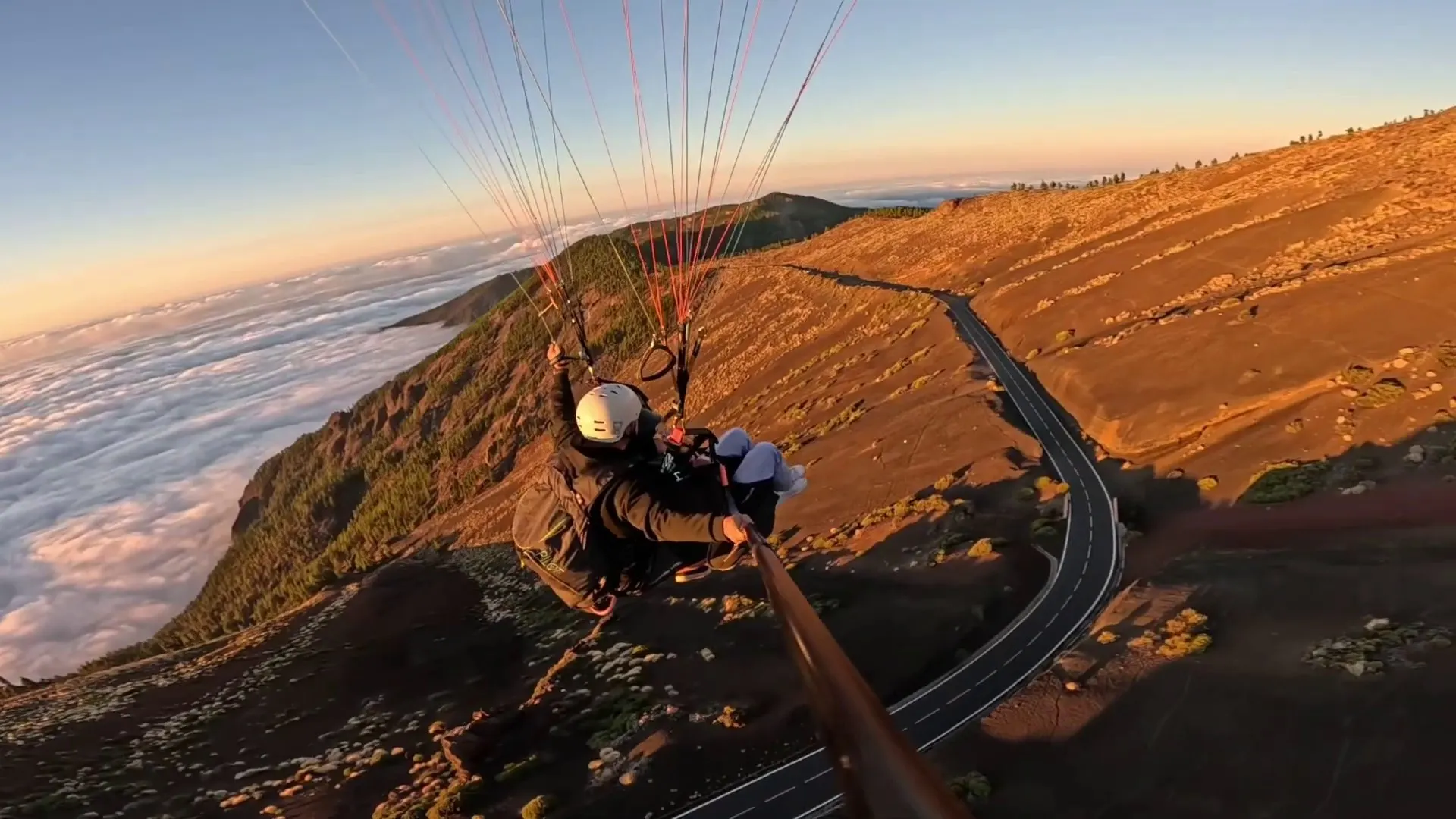 A tandem paraglider over the slopes above the launch, with a mountain road below
