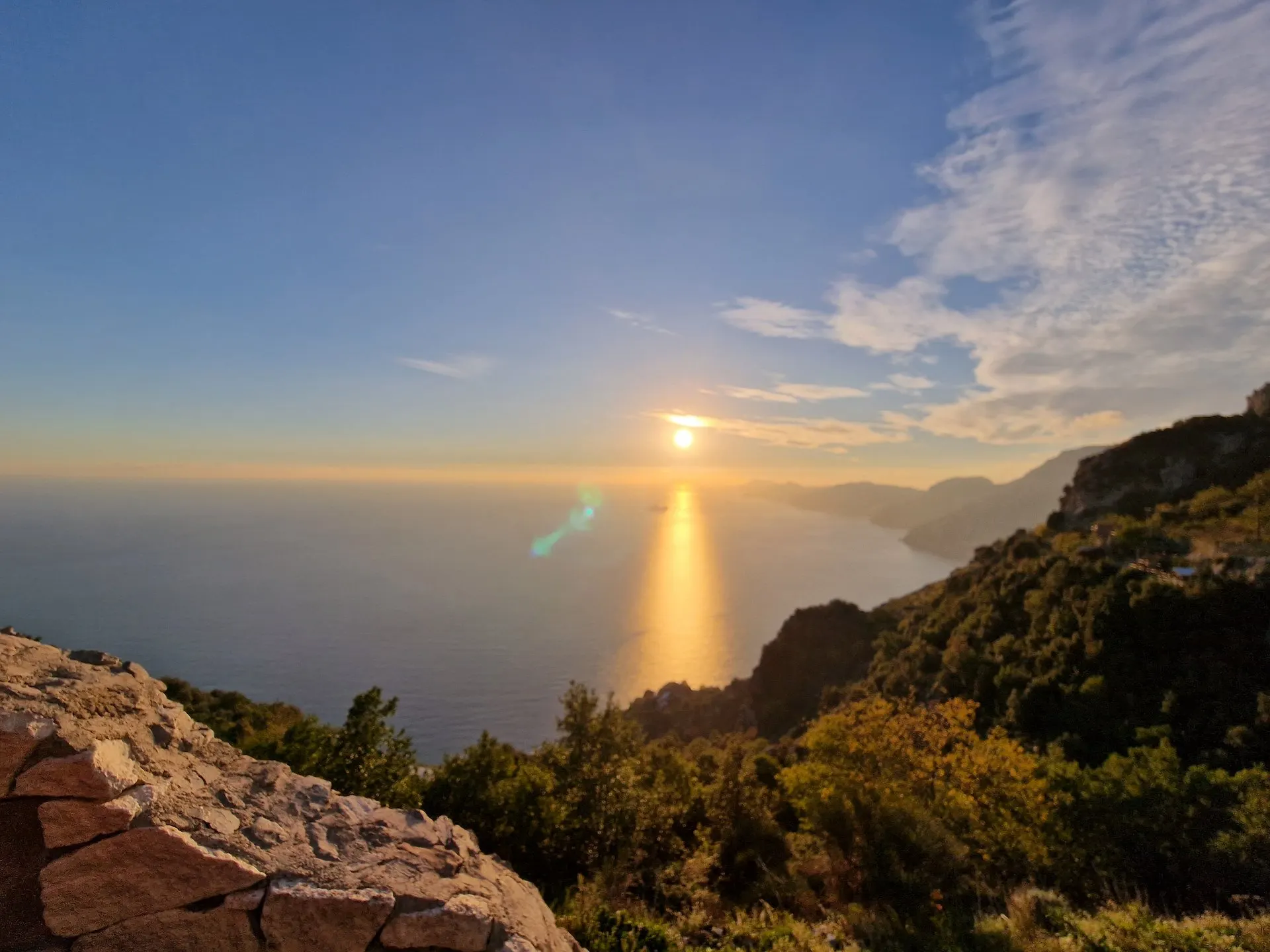 The sun low over the sea, seen from a stone ledge on the mountainside above the coast