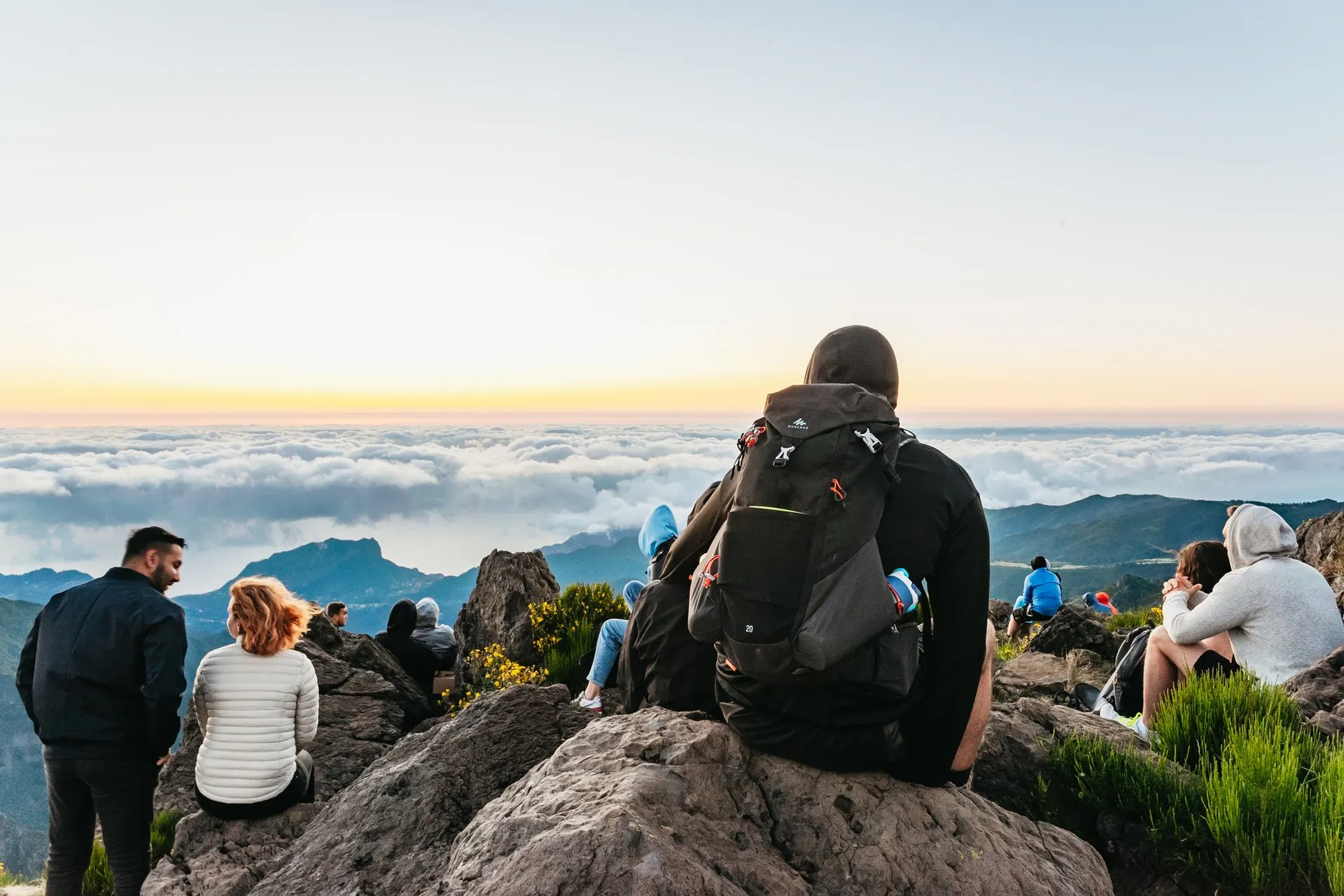 Walkers in warm jackets sitting on the rocks of a mountain top at dawn, looking out over a sea of cloud with green ridges below