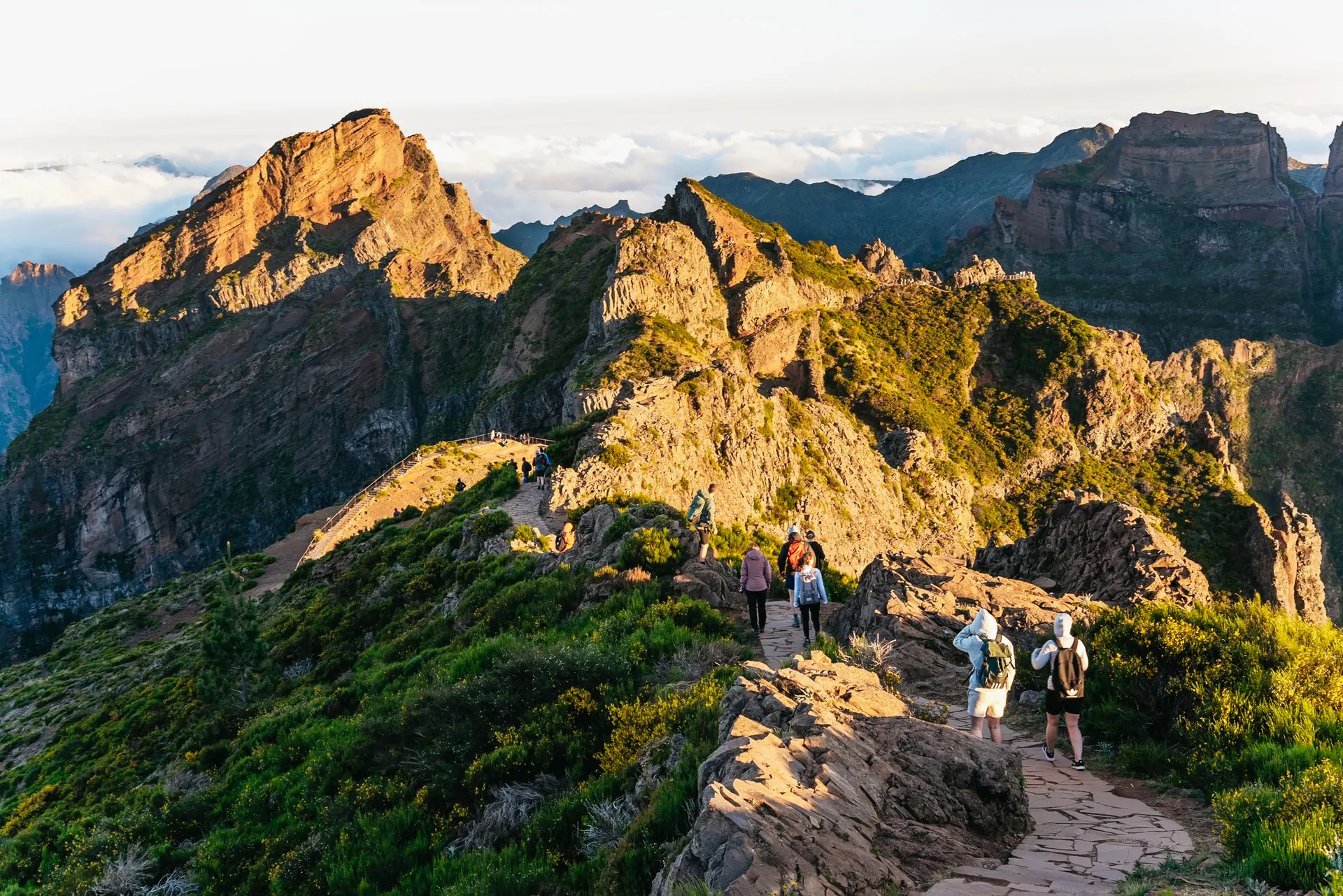 Walkers on a paved path leading from a mountain top towards a railed viewpoint, with sunlit volcanic peaks and cloud beyond
