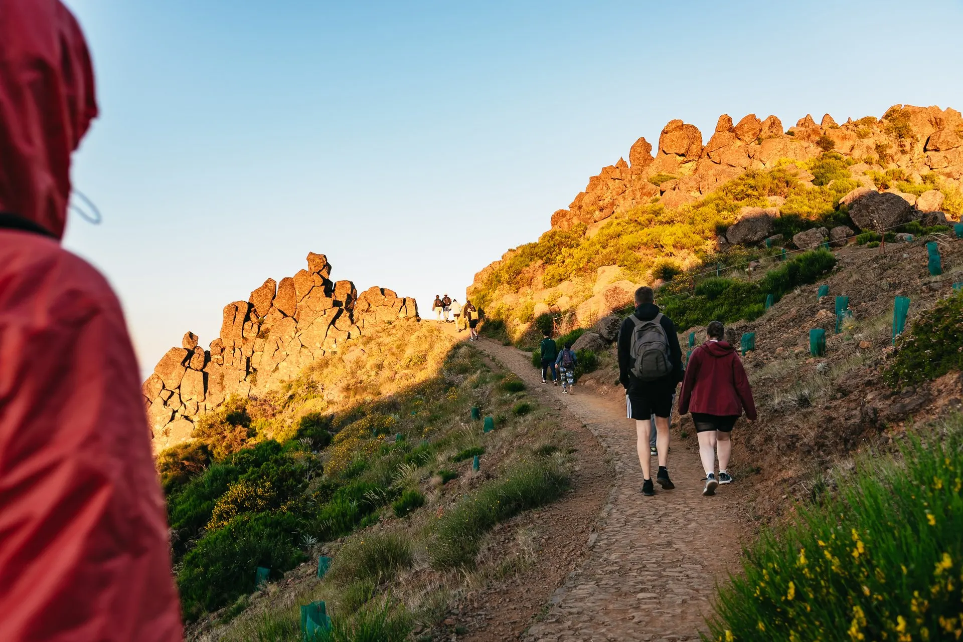 Walkers on a cobbled mountain path below sunlit rock outcrops, under a clear early morning sky