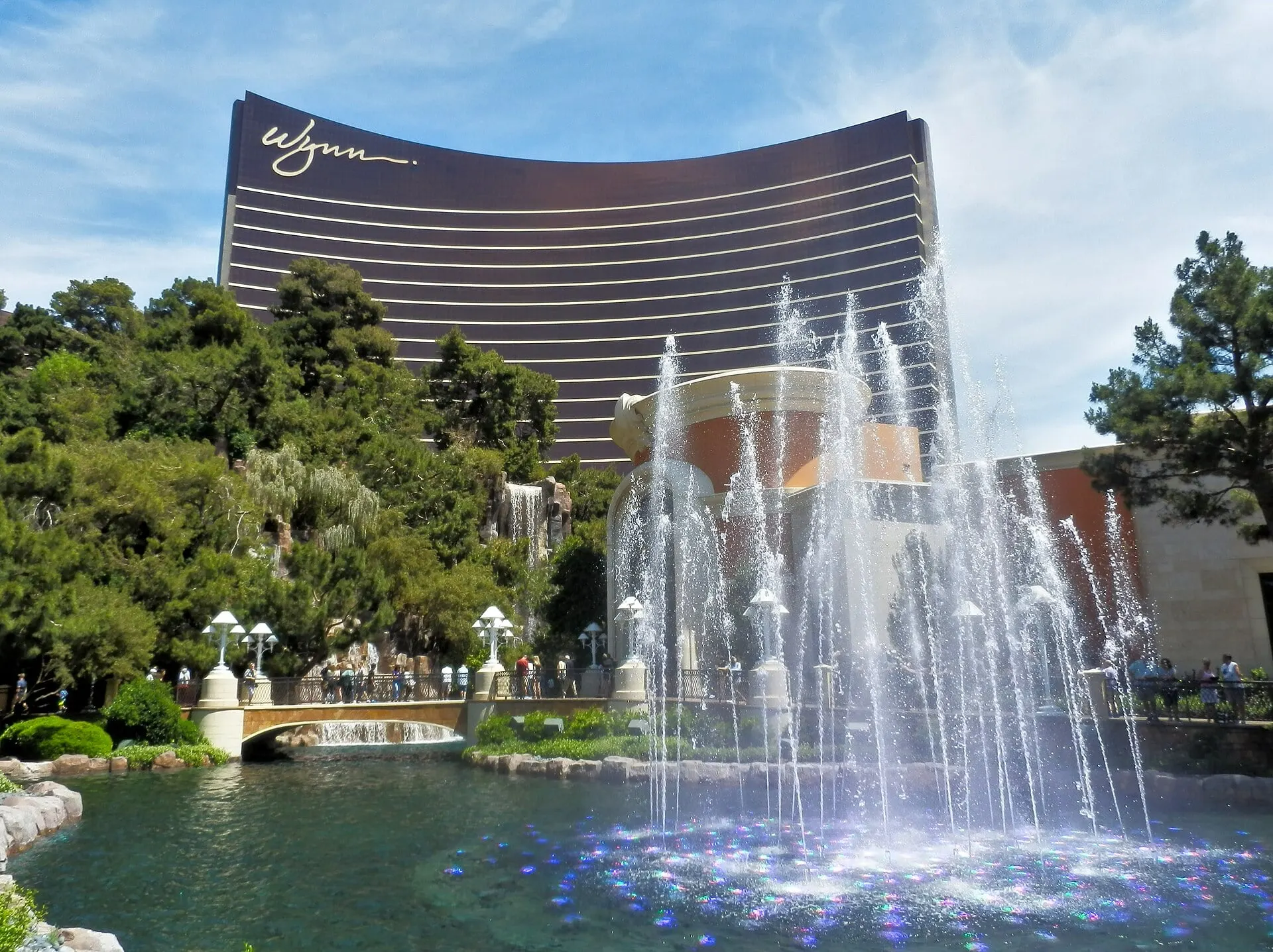 湖の噴水と木々の向こうにそびえる、Wynn Las Vegasの曲線を描くブロンズ色のタワー