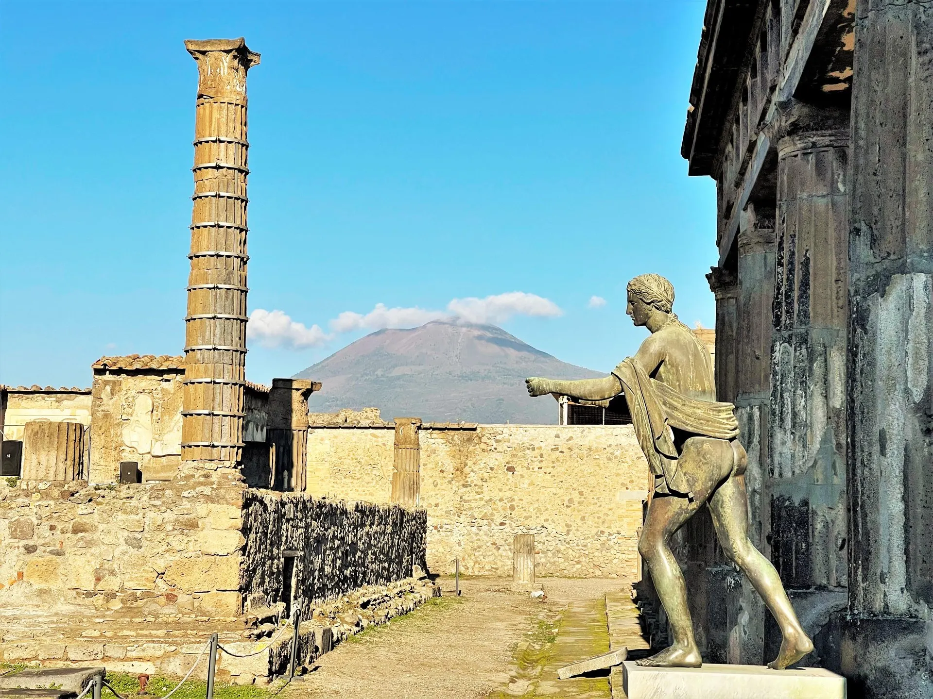 The bronze statue of Apollo beside the Temple of Apollo at Pompeii, with a lone column on the left and Vesuvius under a clear blue sky behind