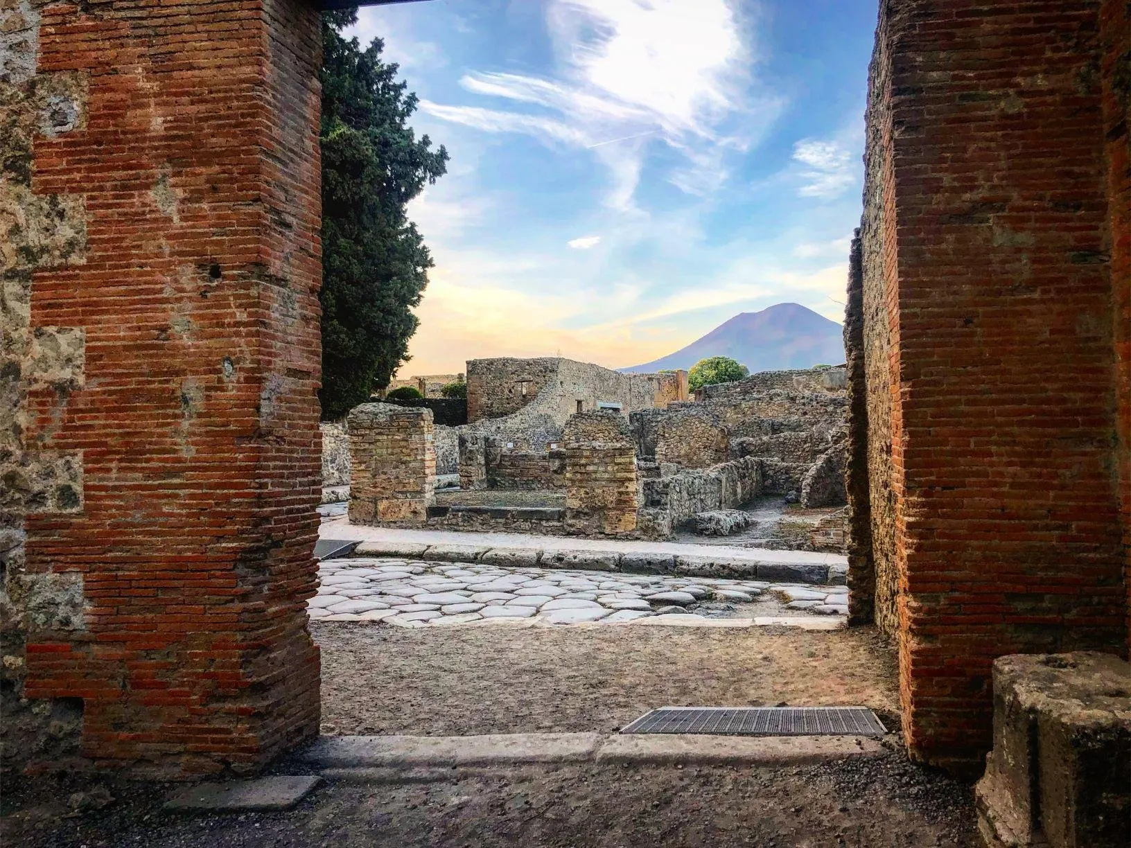 A paved Roman street at Pompeii framed by a brick doorway, with ruined houses beyond and the cone of Vesuvius on the horizon