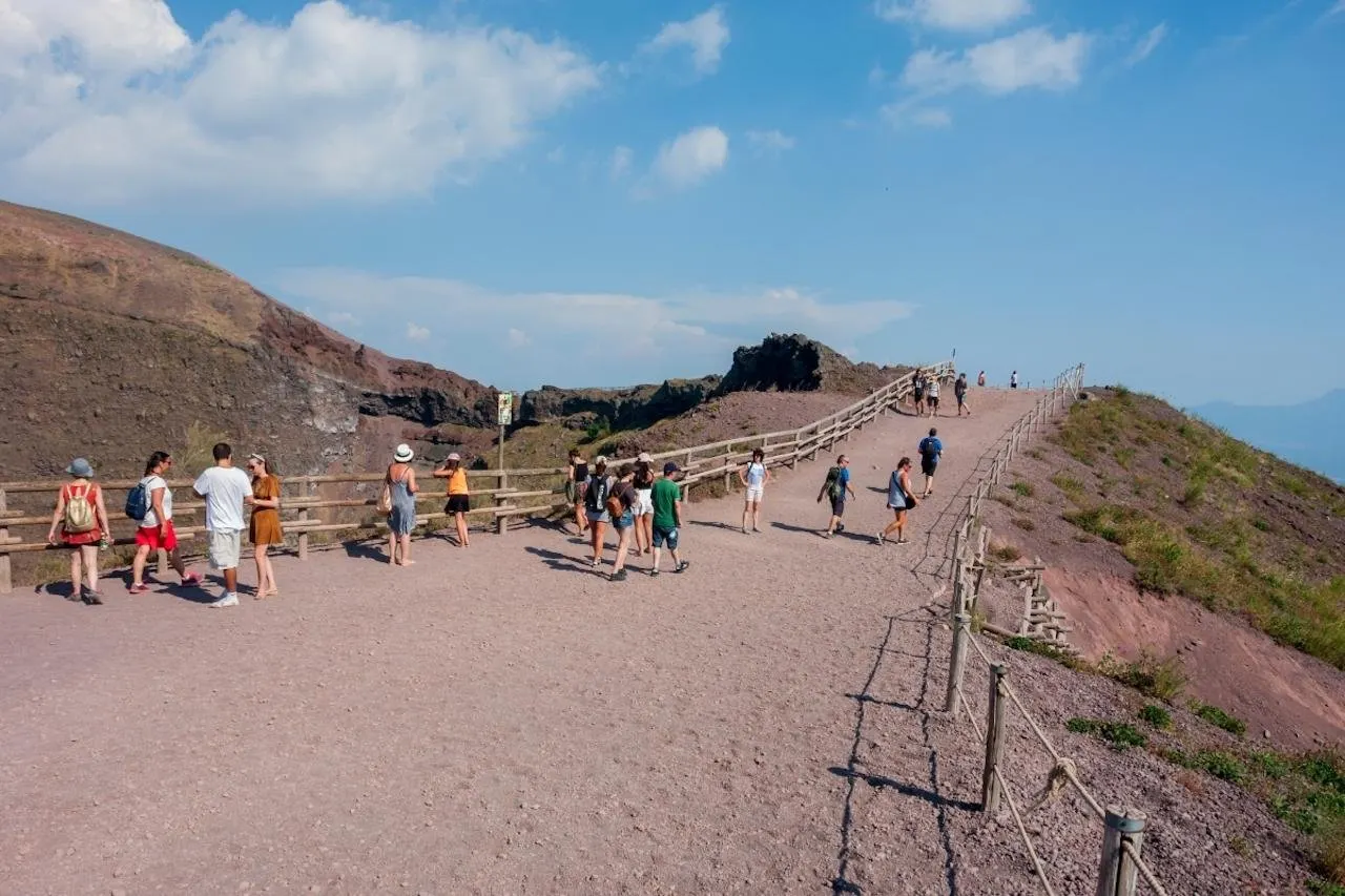 Walkers on the fenced gravel path along the rim of the Vesuvius crater under a blue sky