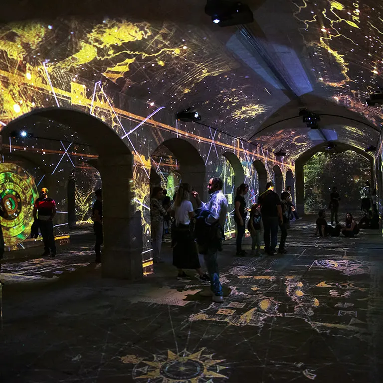 Visitors standing under the stone vaults of the Alfândega furnaces while coloured projections cover the walls, ceiling and floor