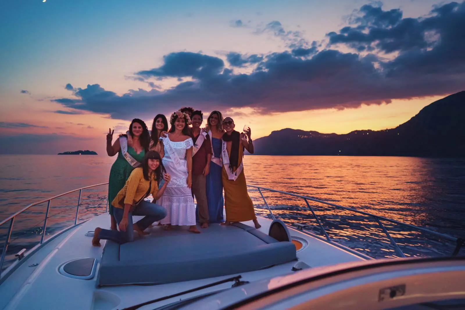 A group of friends on the foredeck of a motor yacht at sunset, with the coast and a small island behind them