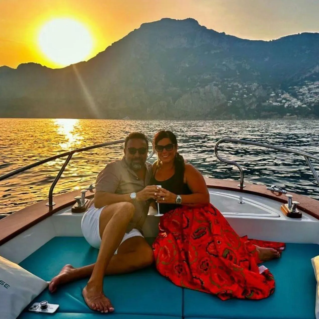 A couple with glasses of prosecco on the bow of a boat, the sun low over the mountains behind them