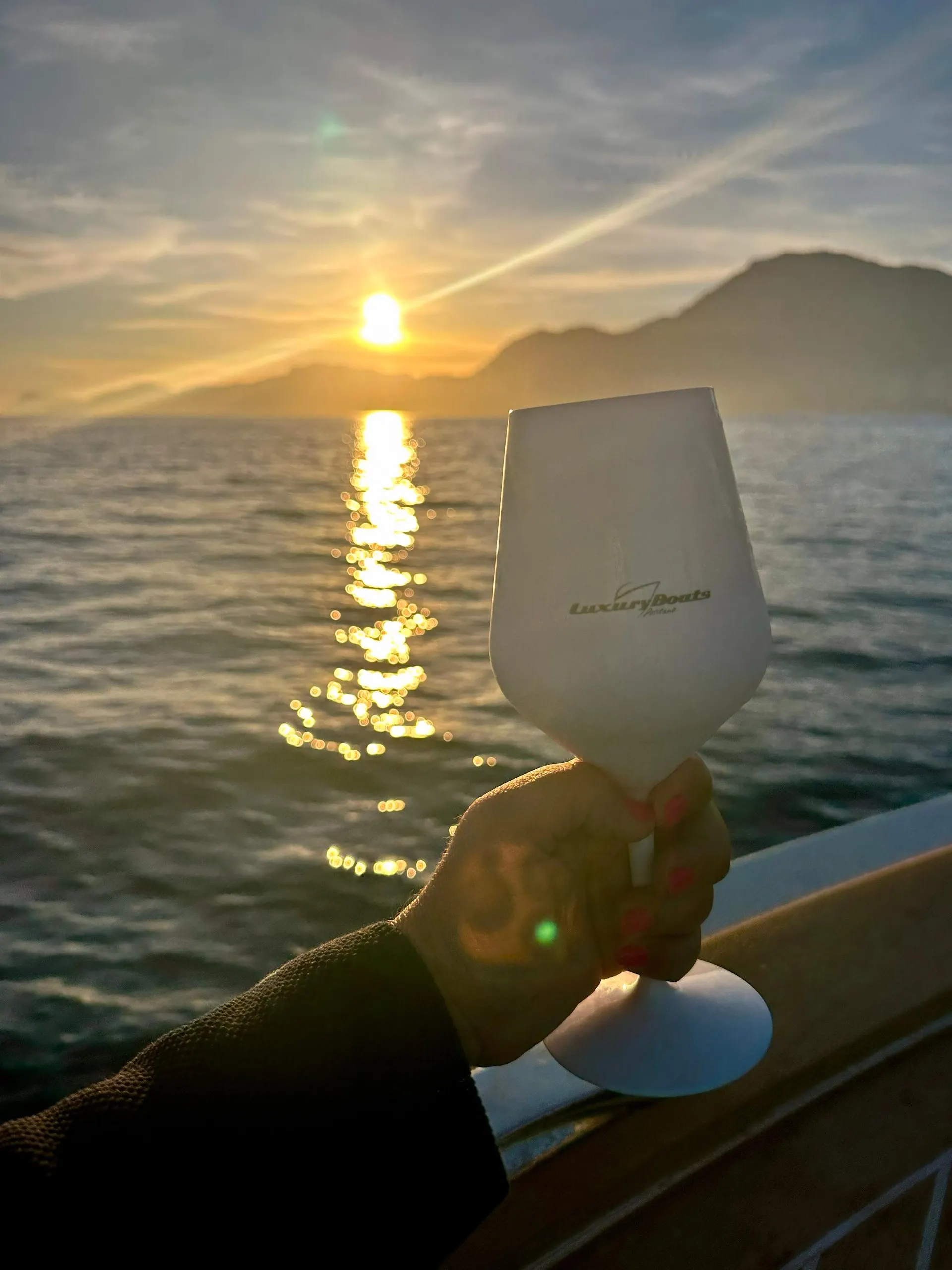 A glass of prosecco held up against the setting sun over the sea, from the deck of a boat