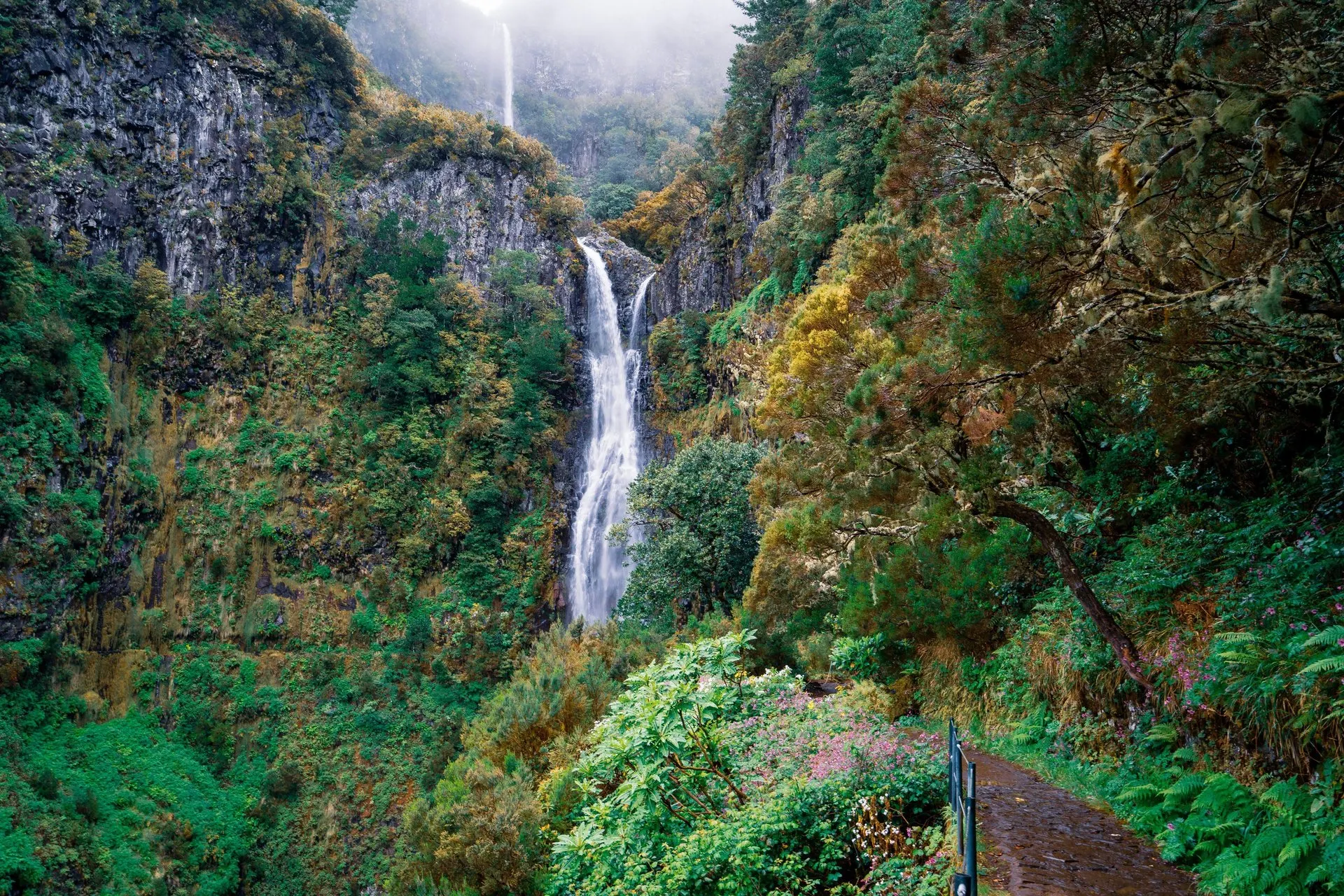 A tall waterfall falling down a forested cliff in the Rabaçal valley, with a railed path in the foreground