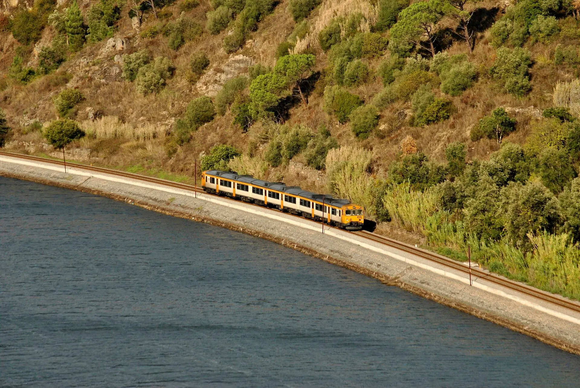Un tren InterRegional de CP sigue la curva del Duero sobre un muro junto al río, bajo una ladera cubierta de matorral.