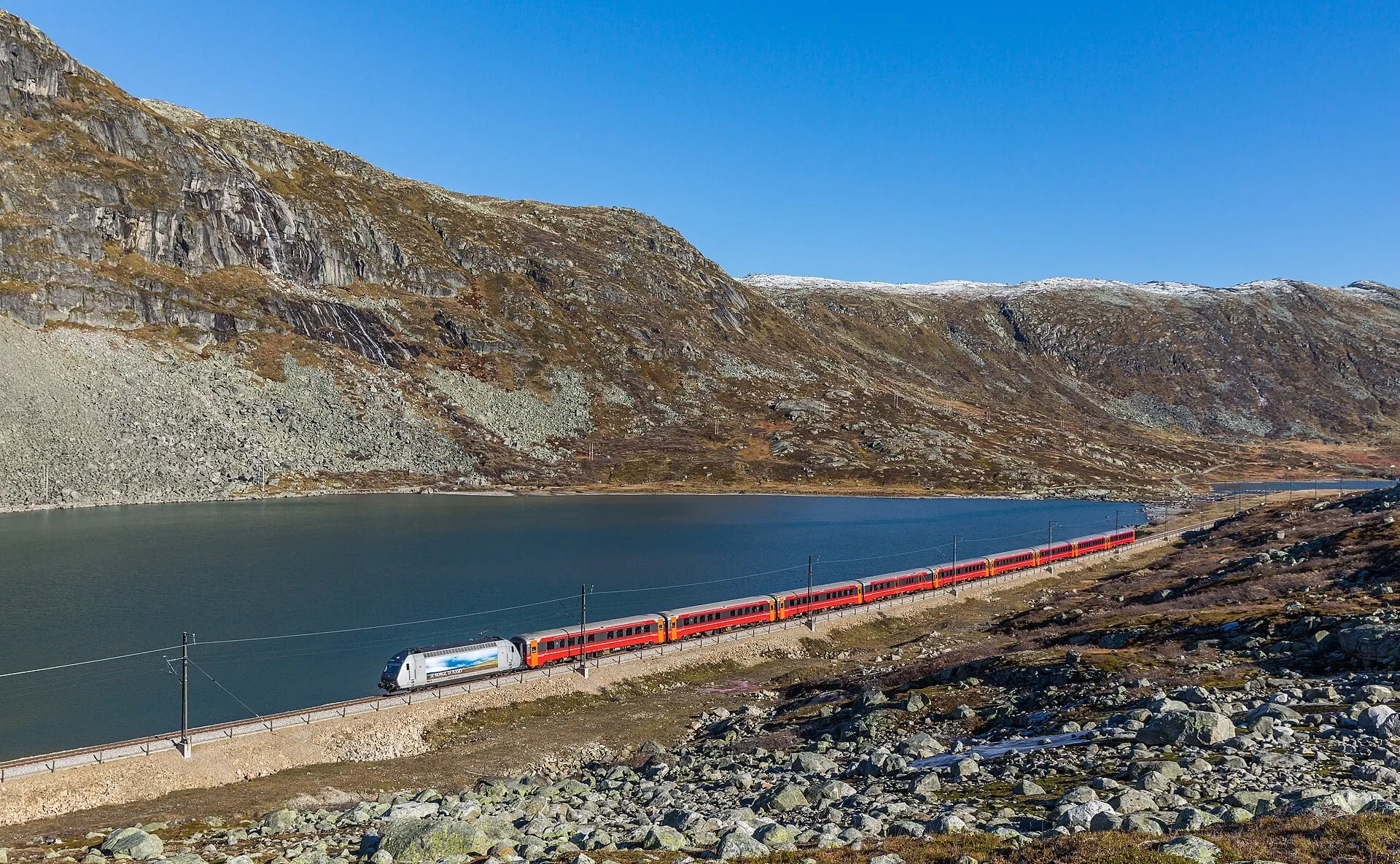 An Oslo to Bergen train passes the lake Nedre Storurdevatnet on Hardangervidda under a clear sky.