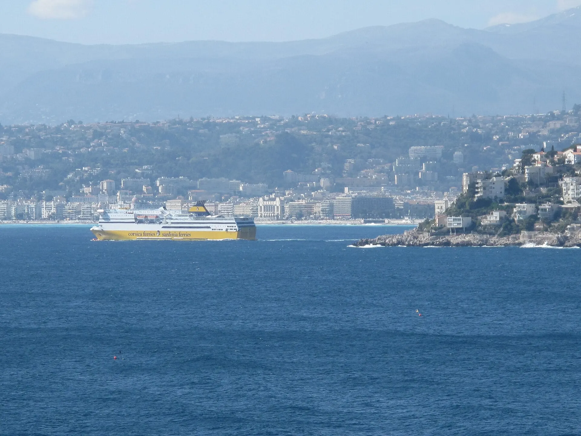 A yellow Corsica Ferries ship sailing out from Nice across the blue bay, with the city and the hills behind