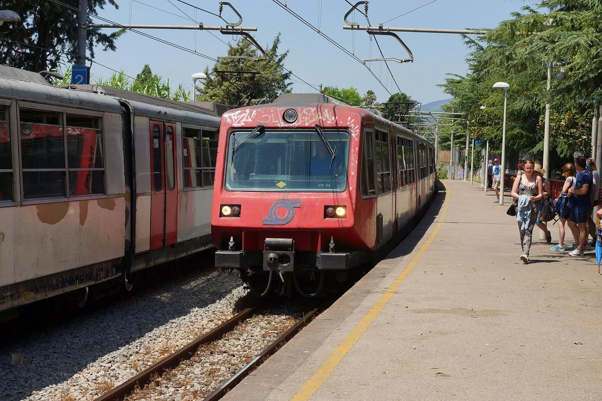 A red Circumvesuviana train at Pompei Scavi station, with passengers on the platform