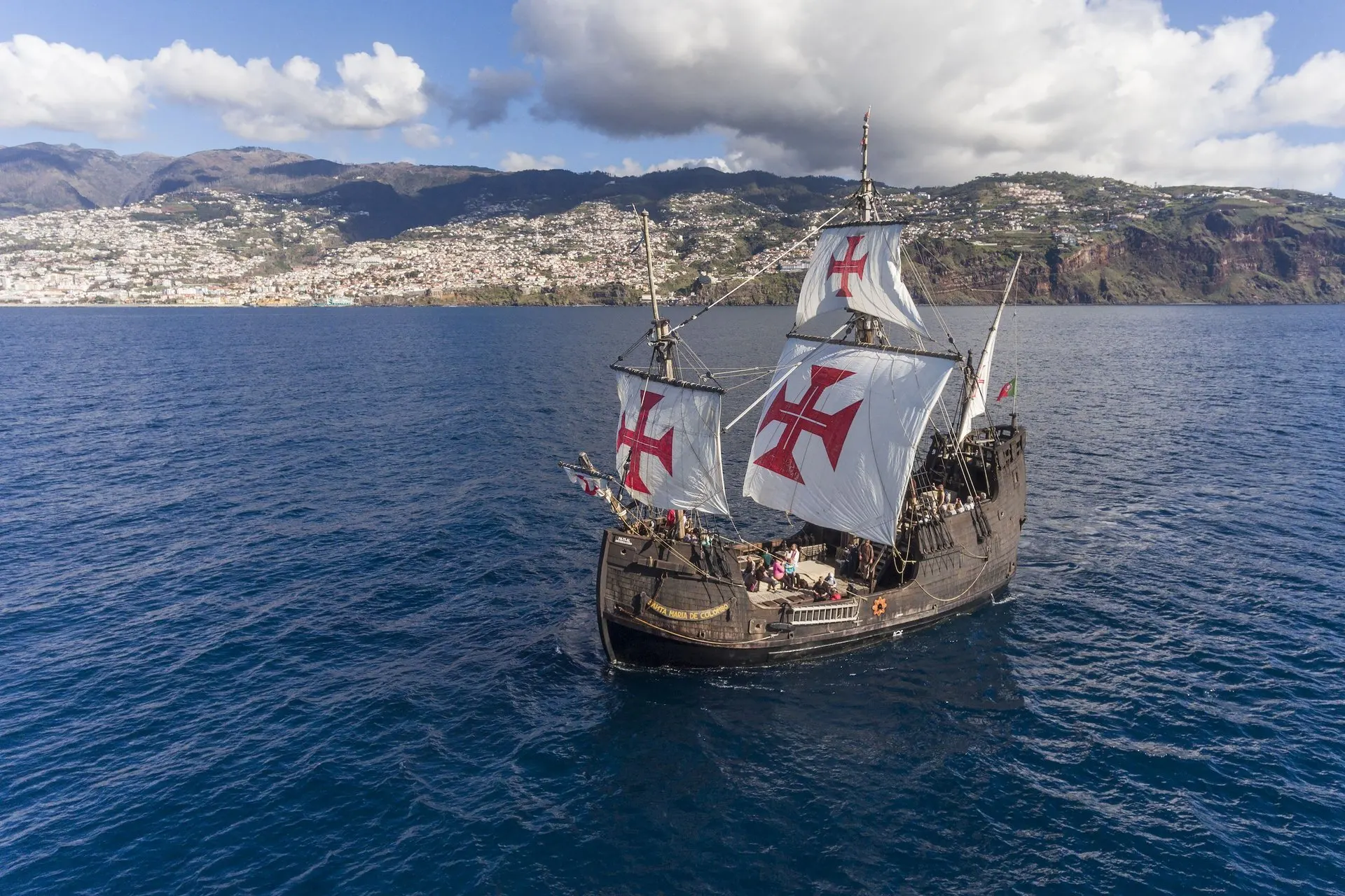 The Santa Maria de Colombo under sail off Funchal, red crosses on her white sails, with the city and its hills behind