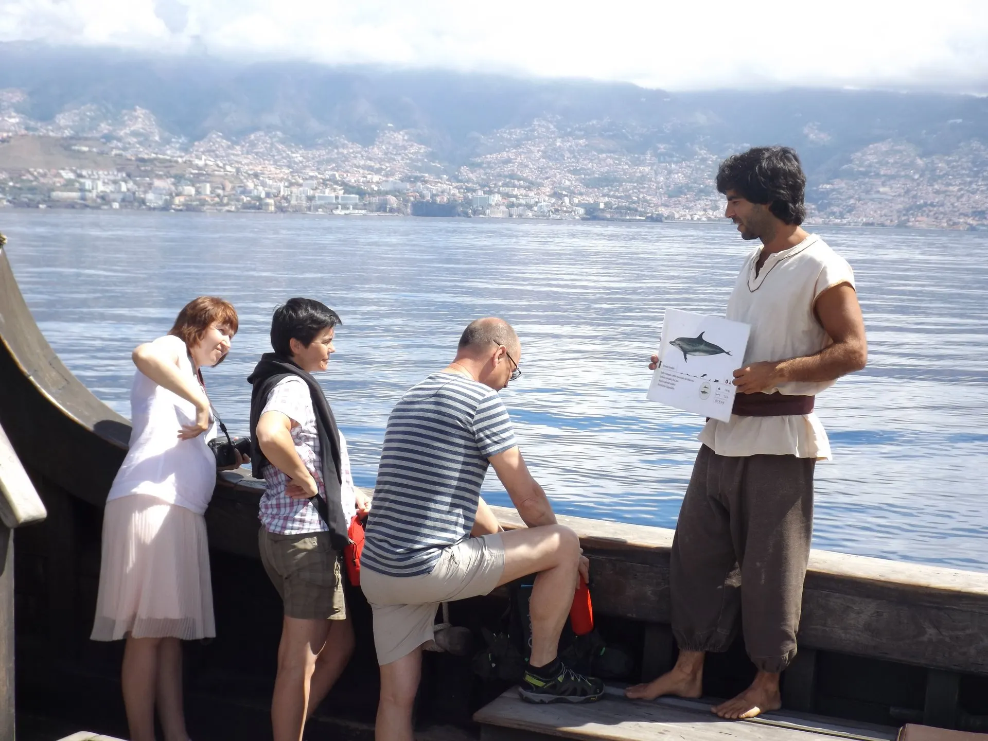A crew member in period dress shows passengers a dolphin identification card at the rail, with Funchal behind