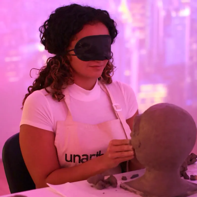 A blindfolded woman in an apron presses clay onto the face of a sculpted head on the table in front of her