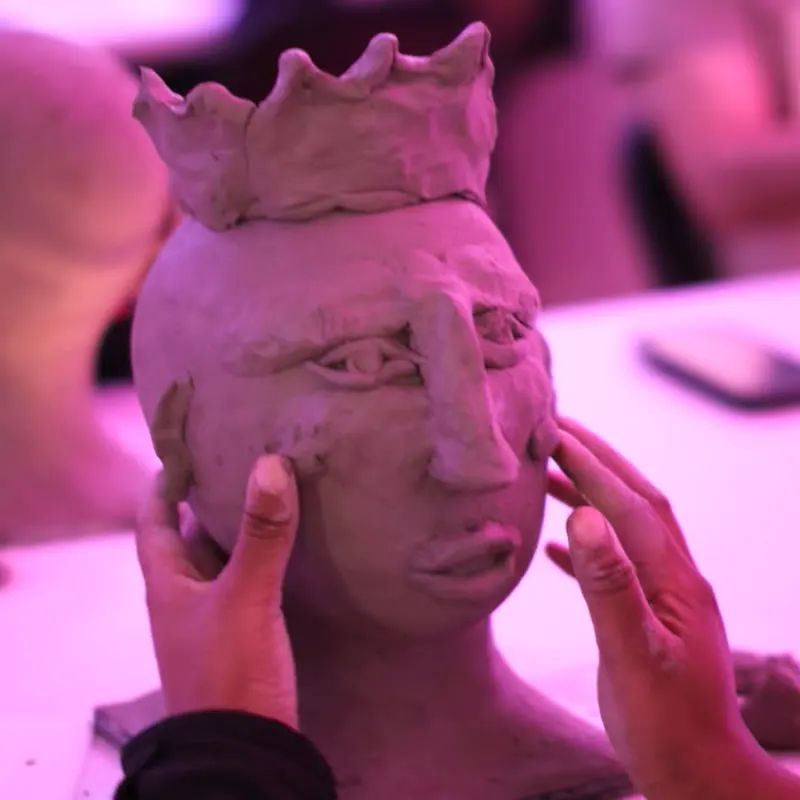 Close view of two hands shaping the eyes and nose of a clay head wearing a clay crown