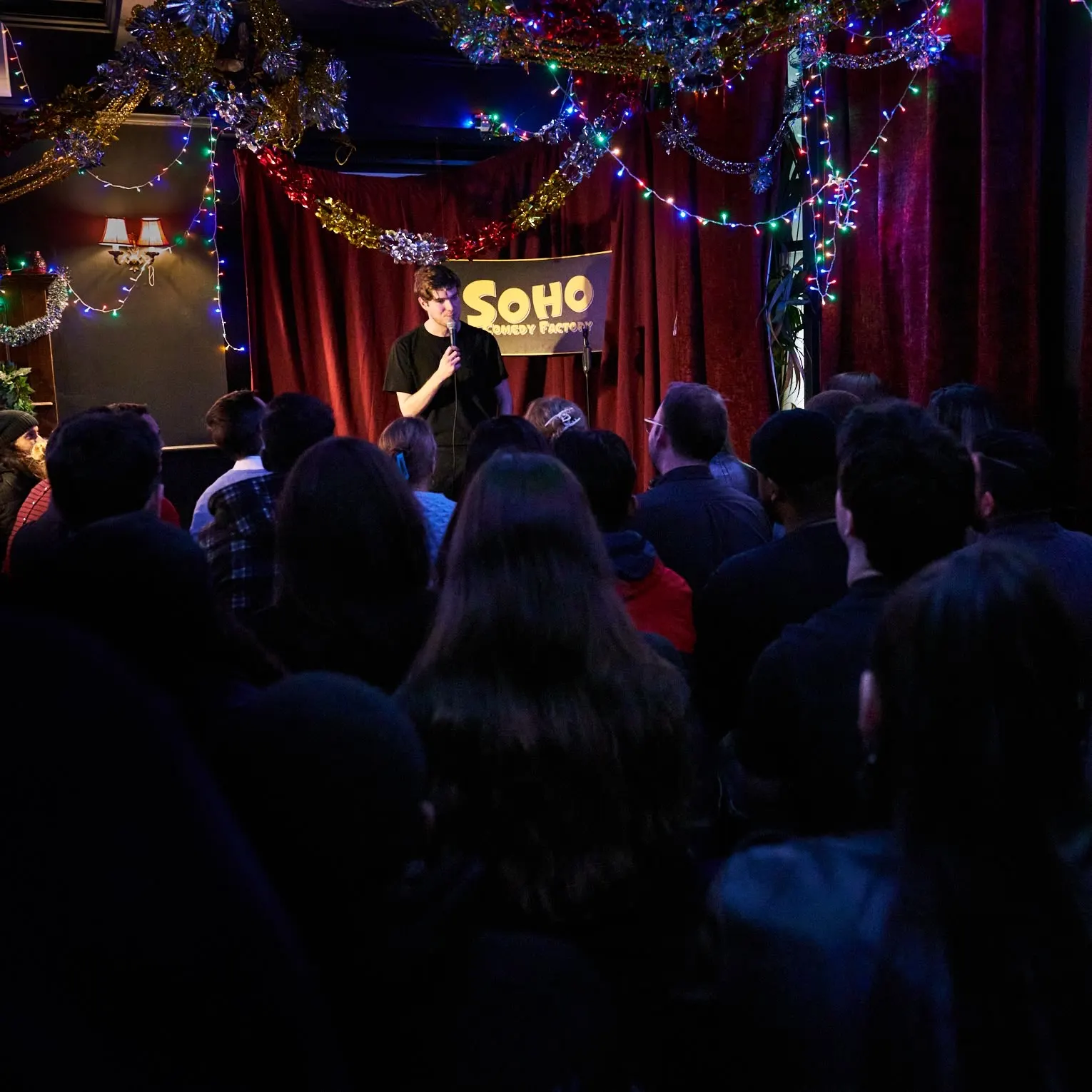 満員の客席越しに見る、赤いカーテンとSoho Comedy Factoryの看板を背にした小さなステージでマイクを持つコメディアン。頭上にはモールと電飾