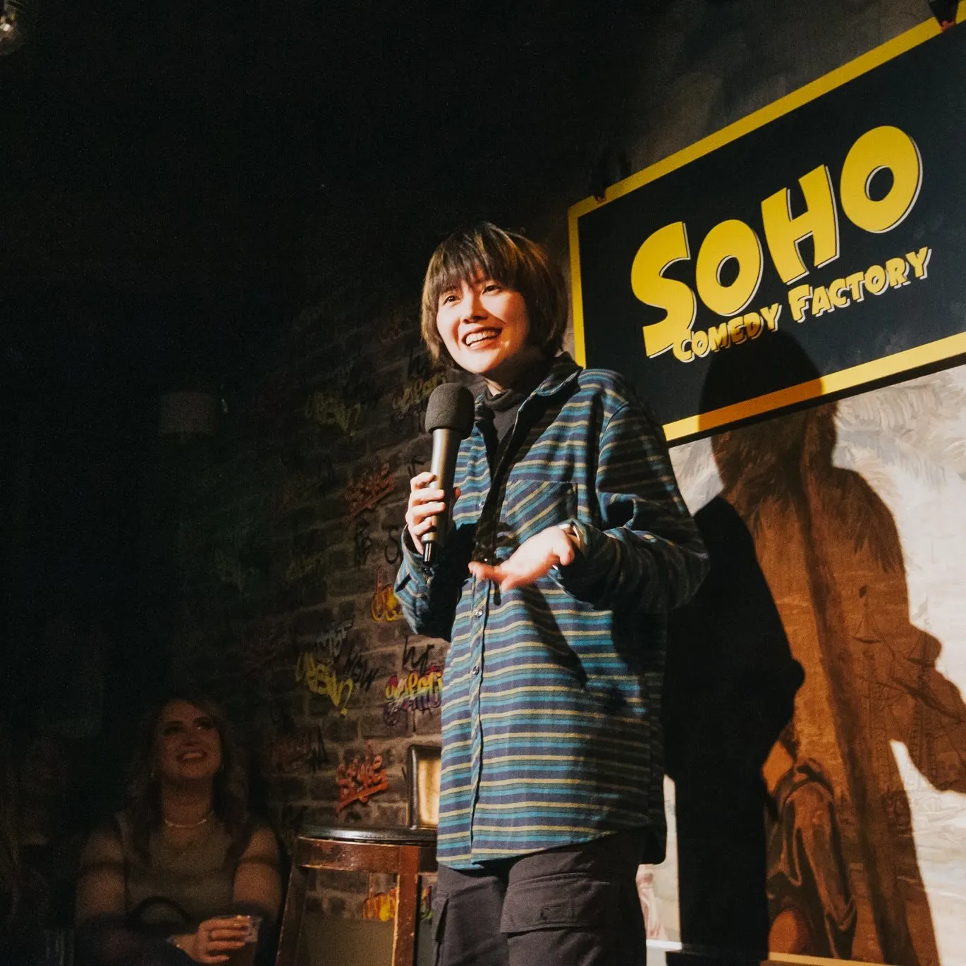 A comedian in a striped shirt smiles into a microphone in front of a brick wall and a yellow Soho Comedy Factory sign