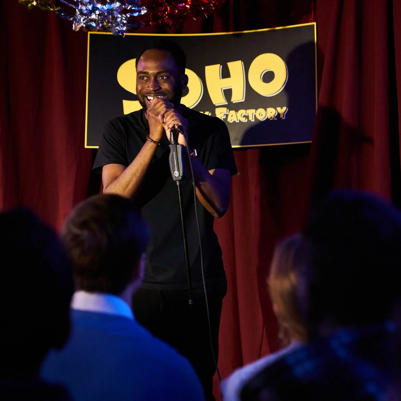 A comedian in a black T-shirt laughs at a microphone stand in front of a red curtain, with audience heads in the foreground