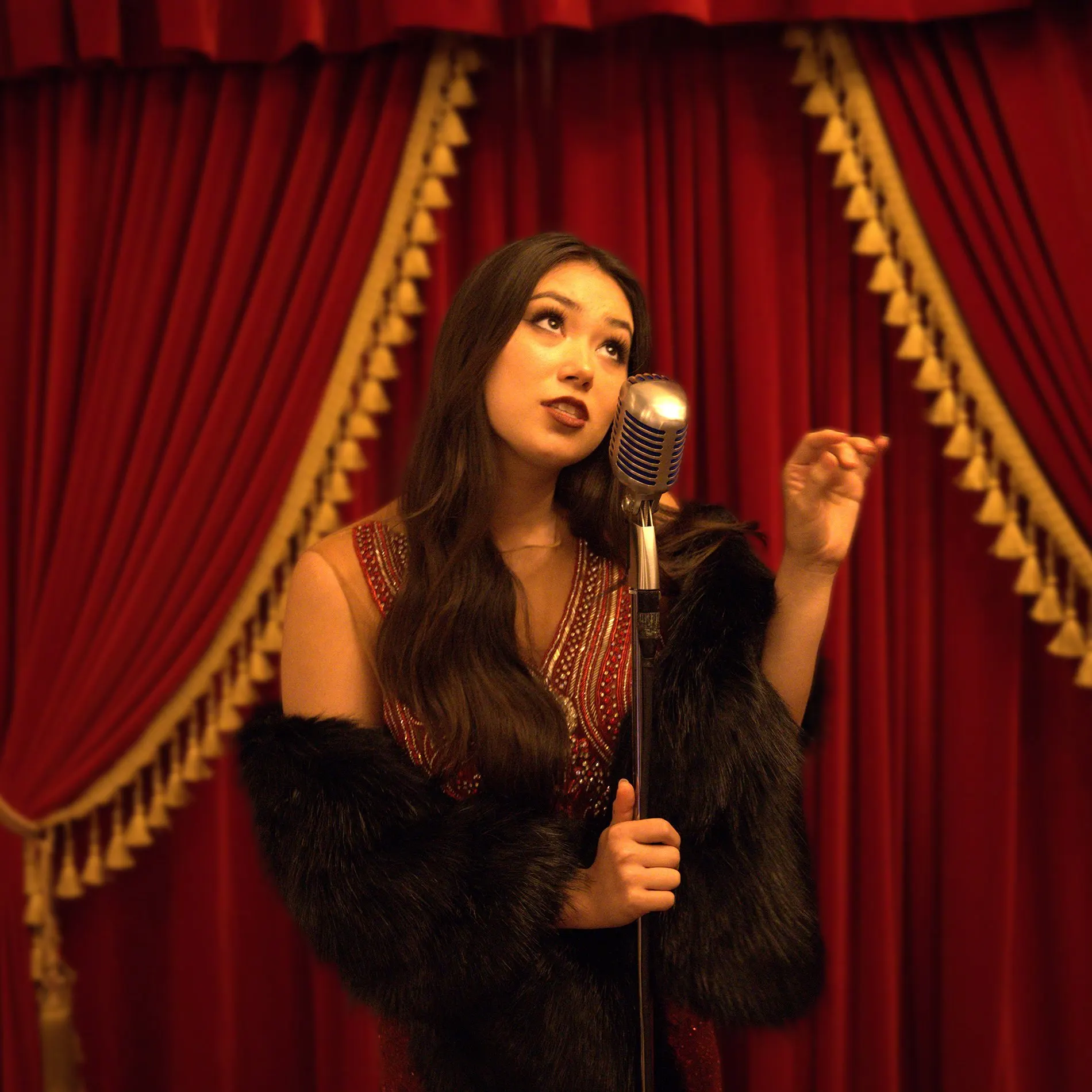 A singer in a beaded red dress and black fur wrap sings into a vintage microphone in front of red velvet curtains with gold tassels