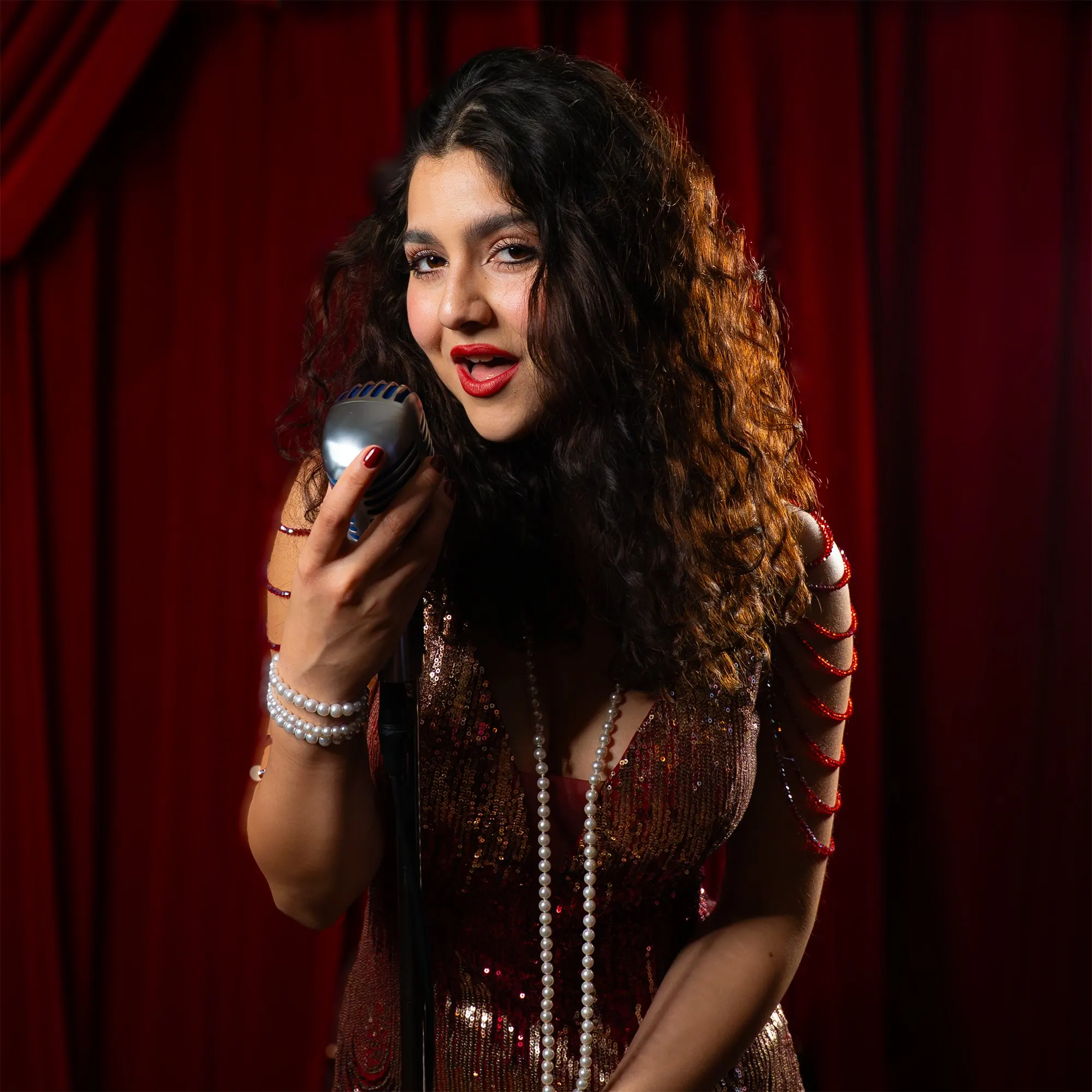 A singer in a sequinned 1920s dress and long pearls holds a vintage microphone against a red velvet curtain