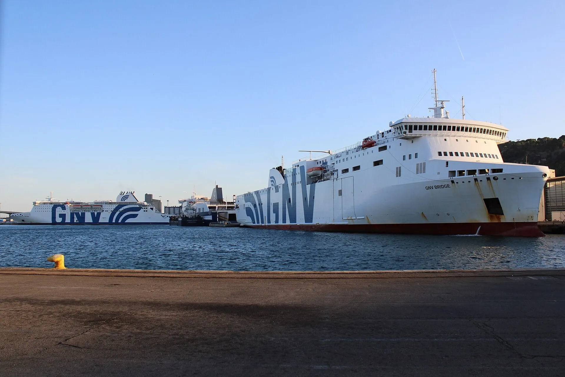GNV ferries moored at the Moll de Sant Bertran in the port of Barcelona, below the green slope of Montjuïc in evening light.